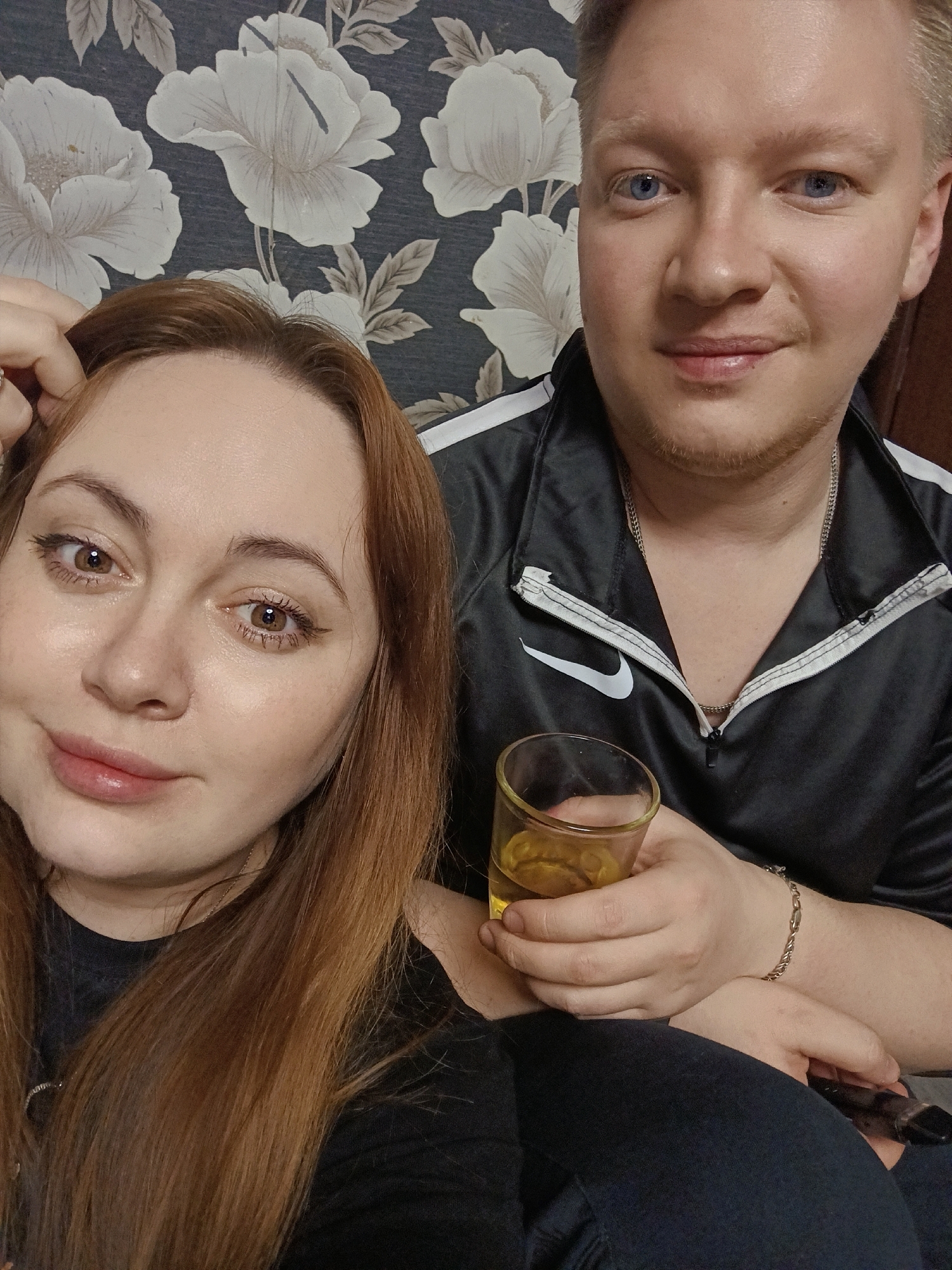 This image depicts two individuals, a female and a male, in a domestic indoor setting within the city of Ольховка, Russia. The background features dark wallpaper with a repeating pattern of large white and light gray floral elements.
The female, positioned on the left and slightly in front, has light skin, long reddish-brown hair, and brown eyes. She is wearing a dark, possibly black, top and has a ring on her left ring finger. Her left hand is raised, with fingers touching the top of her head near her hairline. She is looking directly at the camera with a slight smile.
The male, positioned on the right and slightly behind, has light skin, short blond hair, blue eyes, and light stubble on his chin. He is wearing a black tracksuit-style jacket with white stripes on the shoulders and a white Nike swoosh logo on the chest. A silver chain necklace is visible around his neck, and a silver chain bracelet is on his left wrist. He is holding a small, clear glass containing an amber liquid in his right hand. He is also looking directly at the camera with a slight smile.
The two individuals are seated closely together, with the man's right arm and hand (holding the glass) positioned in front of the woman's right side. A dark-colored remote control is partially visible resting on the man's lap, below the man's hands. The lighting appears to be typical indoor illumination.