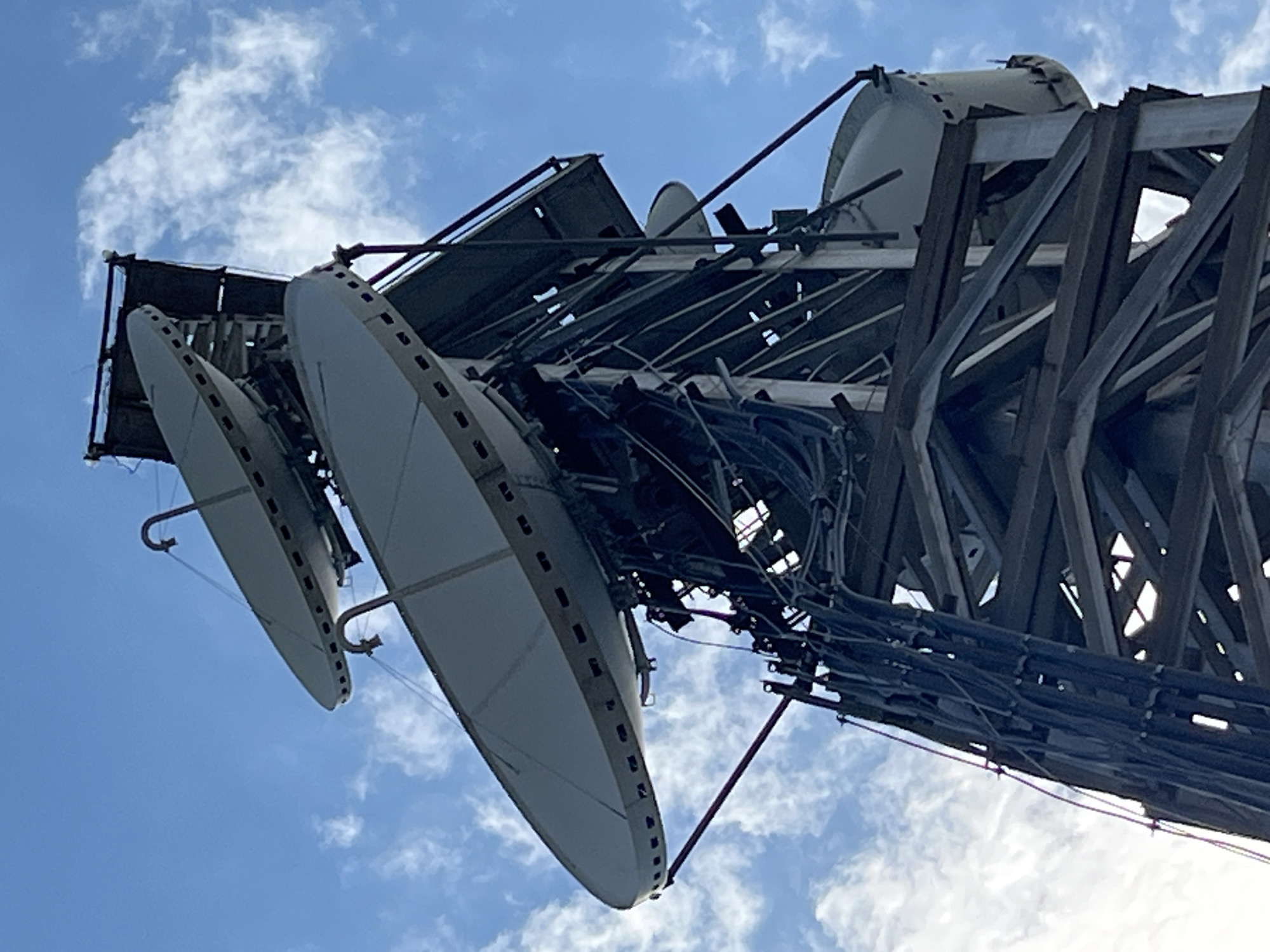 A low-angle, close-up, eye-level, outdoor shot shows a telecommunications tower against a bright blue sky dotted with white, wispy clouds. The tower is constructed of metal beams and supports multiple large, parabolic satellite dishes. Cables run down the side of the tower. The tower appears to be located in a generally clear area, as indicated by the open sky. No people are visible in the image. This telecommunications tower is situated in Cholpon-Ata, Kyrgyzstan.