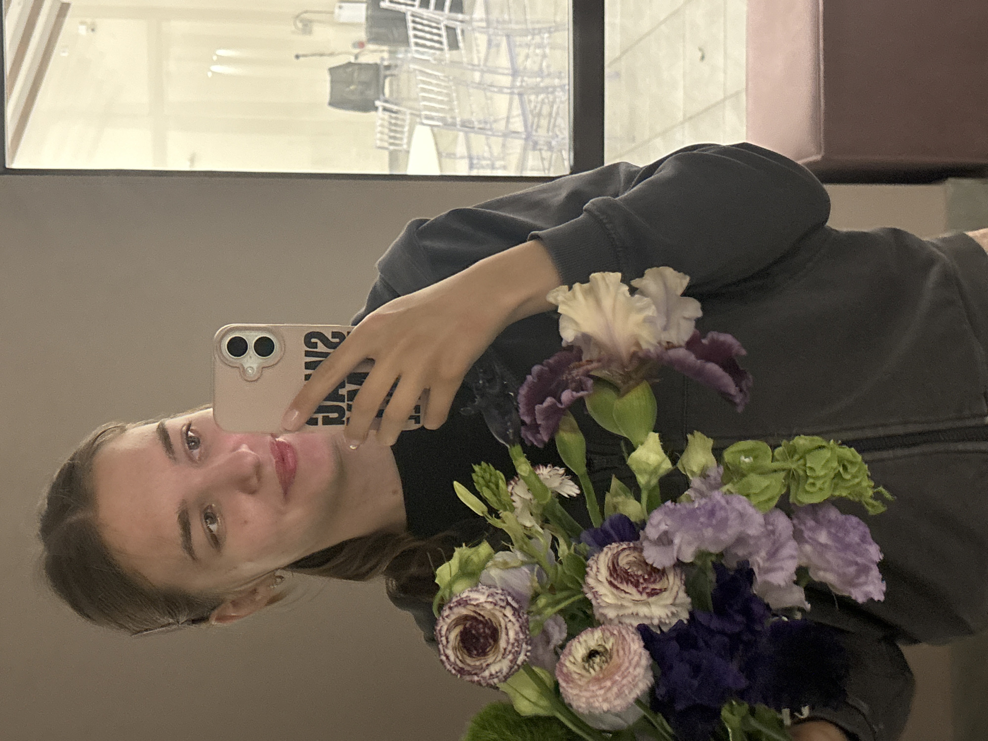 A young woman with light brown hair pulled back in a ponytail is taking a mirror selfie. She is holding a bouquet of purple and pink flowers in her left hand and her smartphone in her right hand. She is wearing a dark gray hooded sweatshirt. The background is a reflection in a mirror, showing a room with tables and chairs, possibly a venue or event space. The setting appears to be indoors with a large window or glass partition on the right side of the image. The location is identified as Nizhniye Mully, Russia.