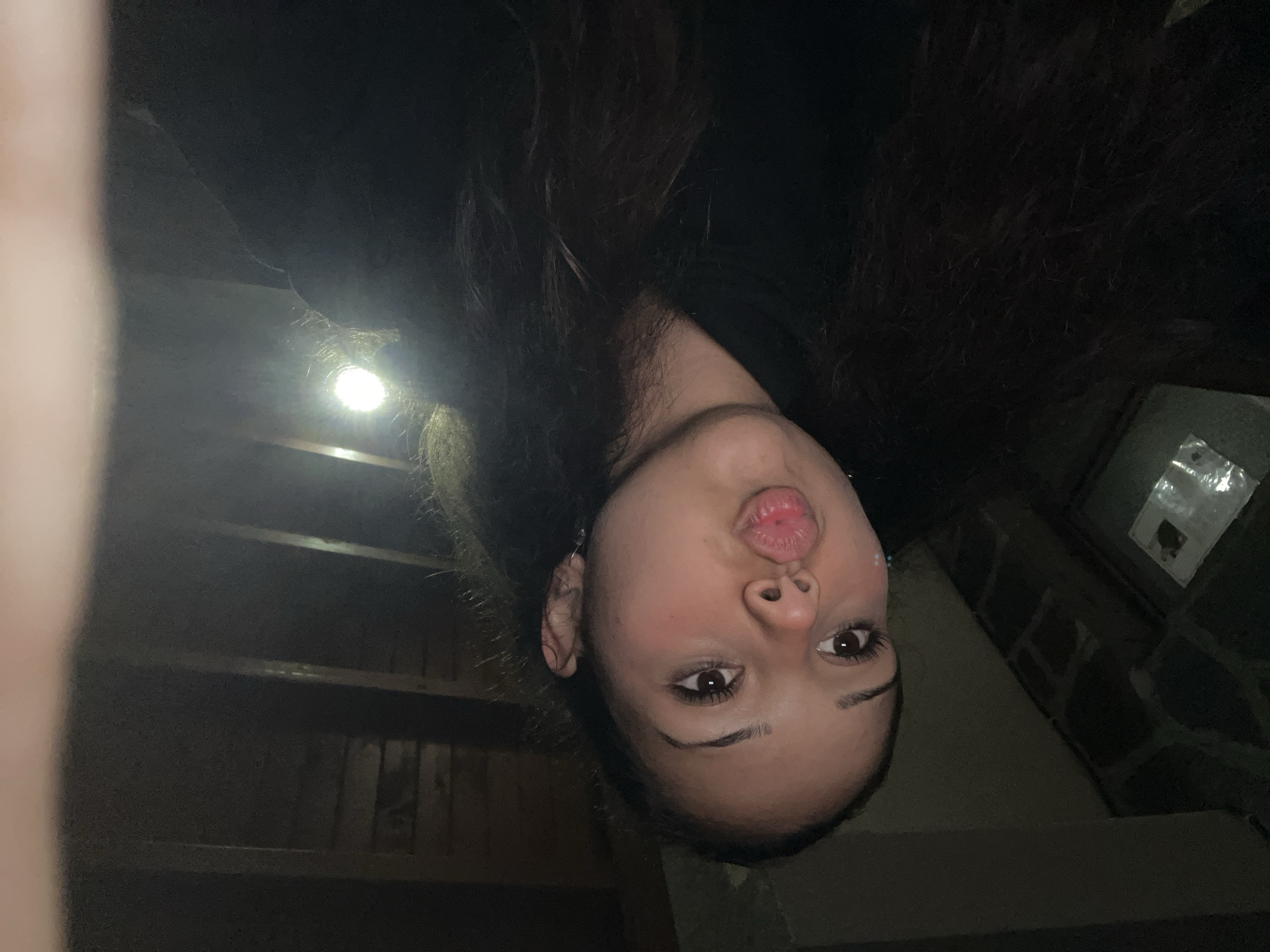 A young woman with dark hair is captured in a close-up selfie. She is wearing a black hoodie and has her lips puckered in a kiss. The background appears to be a dimly lit room with wooden paneling on the ceiling. The lighting suggests it might be nighttime or an indoor setting with artificial light. No specific details about the city of Zlatograd, Bulgaria, are visible in the image.
