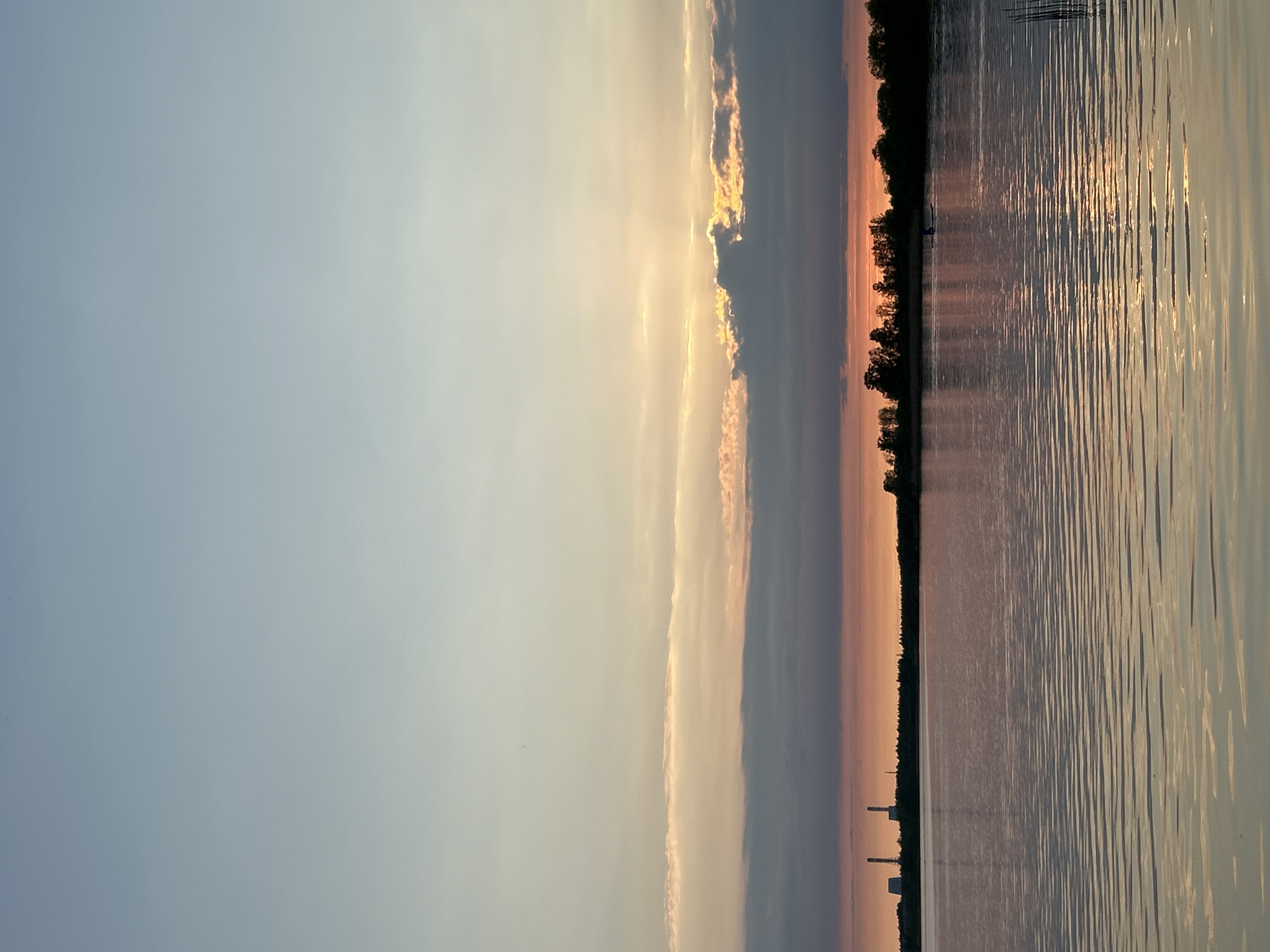 The image captures a sunset over water, likely the Neva River or a nearby body of water in Saint Petersburg, Russia. A pale blue sky transitions into bands of clouds illuminated by the setting sun. The water reflects the golden light, creating vertical streaks and reflections. Along the horizon, a line of trees is visible. The image is taken at an angle. In the bottom center near the tree line some man made structures are visible.