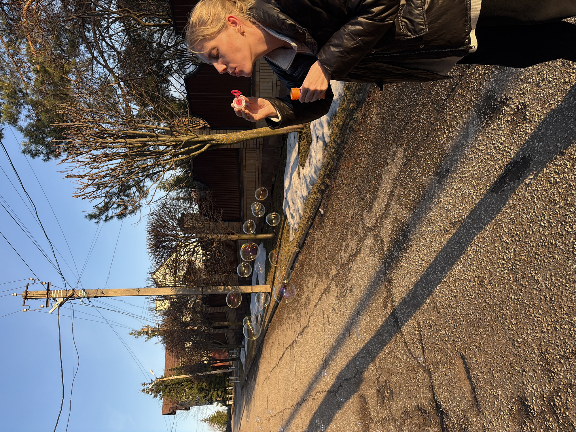 The photograph shows a woman blowing bubbles on a street in Прикамский, Russia. The woman, wearing a dark jacket, is holding a bubble wand and container. Bubbles are floating in the air. The street is asphalt and has patches of shadows on it. In the background are houses, trees, and a utility pole with power lines. There is also a patch of snow on the side of the road. The sky is blue.