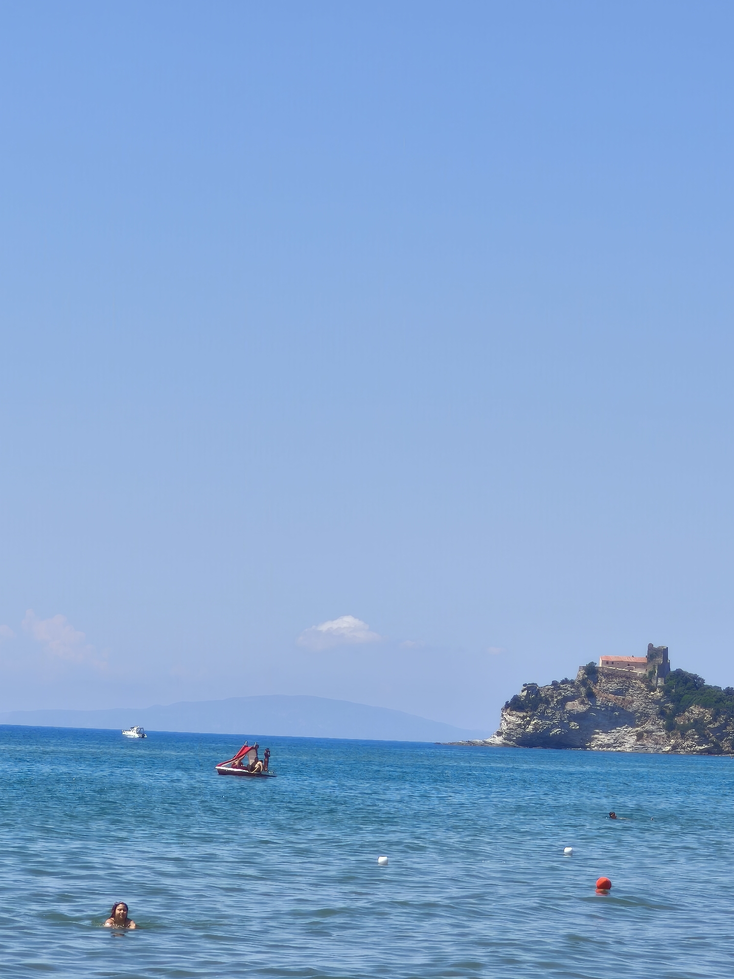 A clear, bright summer day unfolds over the waters near Castiglione della Pescaia, Italy. The azure sky is vast and cloudless, with a few wispy formations in the distance. The deep blue sea, calm and inviting, stretches towards the horizon.
In the foreground, a young person with dark hair is partially submerged in the water, facing forward with a slight smile, wearing headphones. Further out, a small red paddleboat is occupied by at least two individuals, one steering and another standing. A white motorboat is visible in the middle ground.
Dominating the background is a rocky promontory topped by a fortress-like structure with red-tiled roofs. This iconic landmark is situated on the coast, with the rugged cliffs dropping sharply to the sea. The distant coastline is a faint, hazy line of mountains.
Several buoys mark an area of the water, including a bright orange one and two white ones. The overall scene depicts a tranquil day at the beach, with recreational activities taking place in the sea against a backdrop of historical architecture and natural beauty.