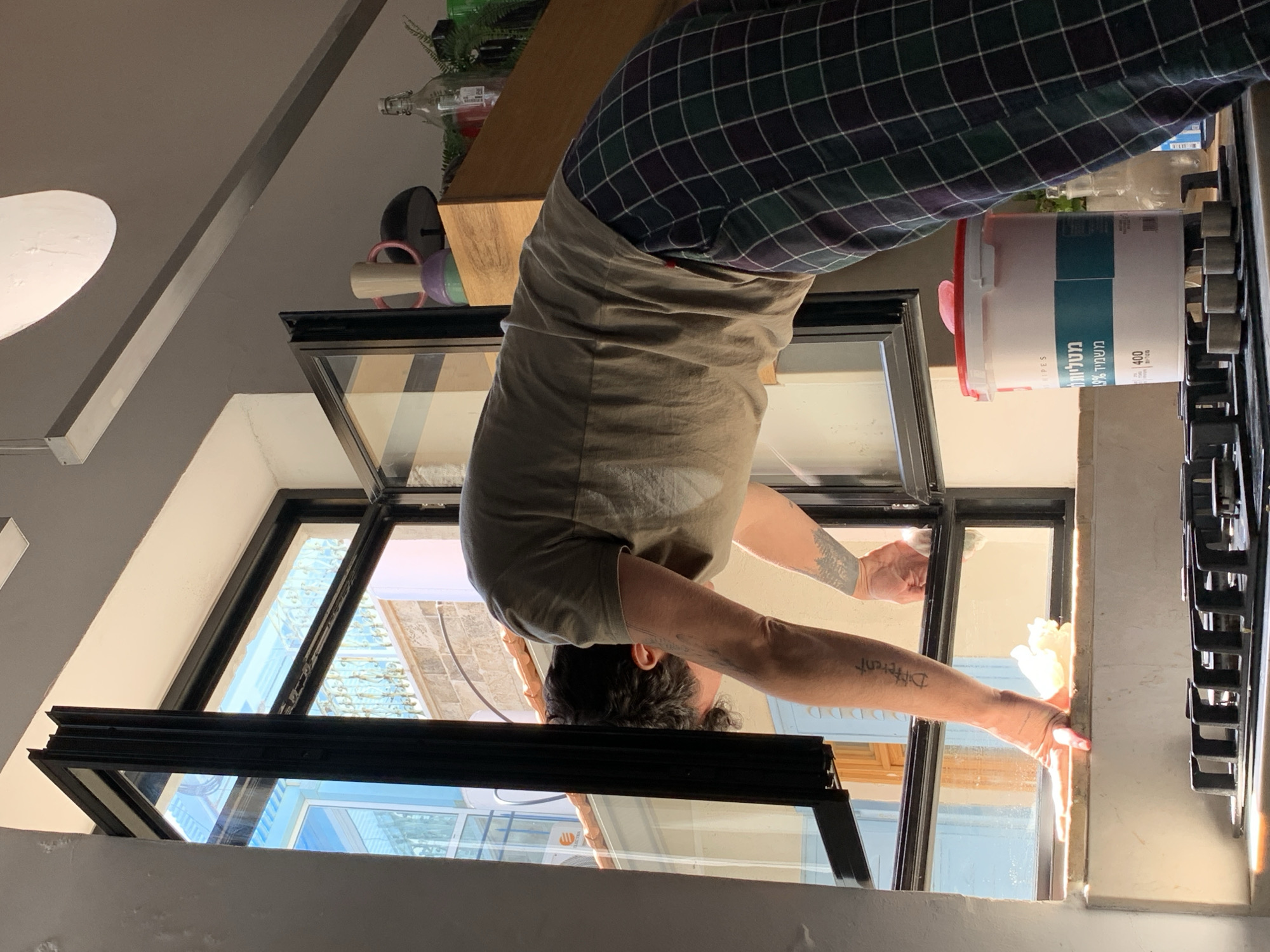 The image depicts a man upside down, leaning through an open window, possibly cleaning it. He is wearing a t-shirt and plaid pajama pants. His arms are extended, and there is a cleaning bucket next to him on what appears to be a kitchen countertop. The window reveals a view of a Tel-Aviv neighborhood, with buildings and balconies visible.