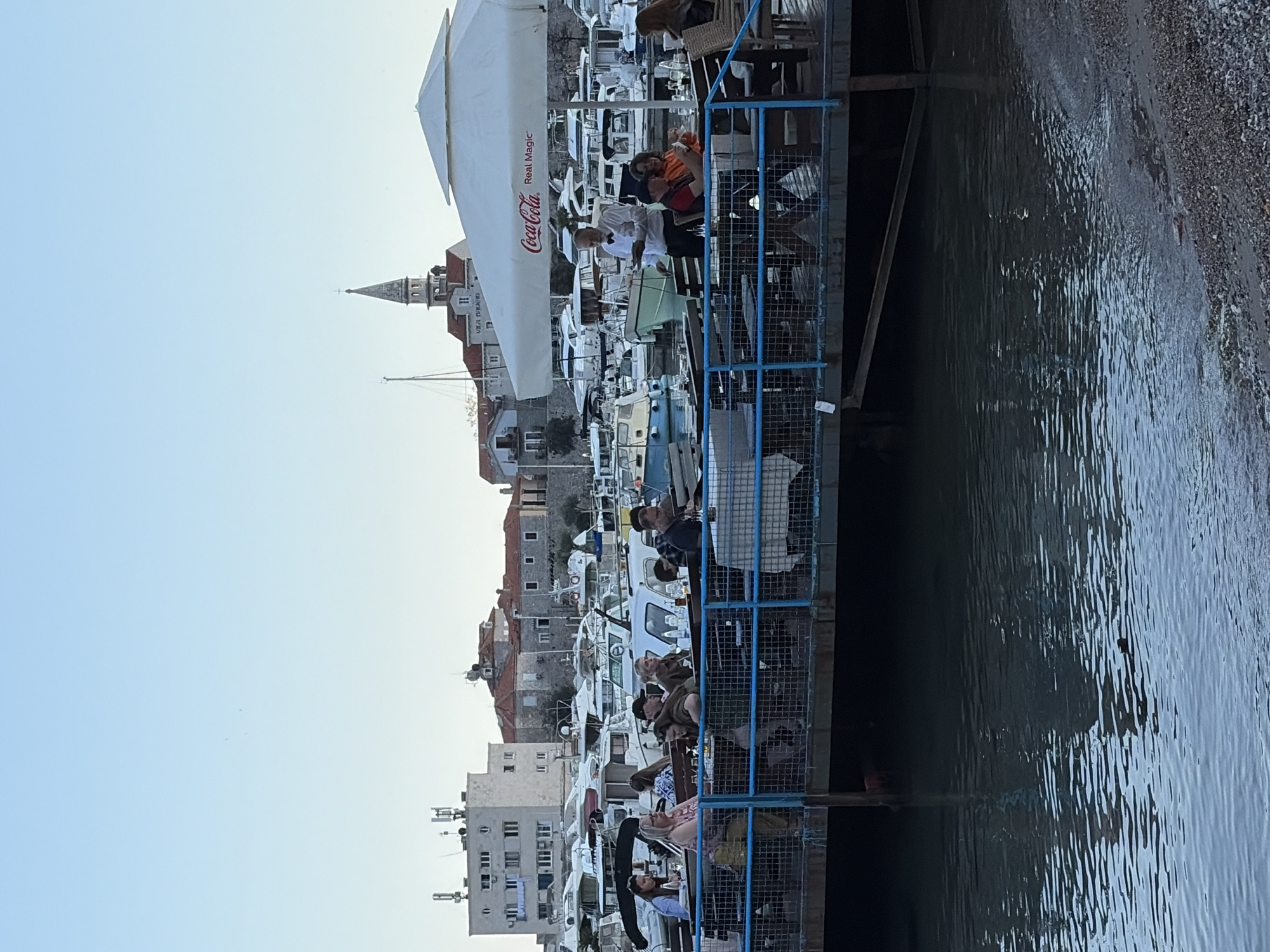 The image depicts a waterfront scene in Budva, Montenegro. A restaurant or bar extends over the water, with a blue metal railing enclosing the seating area. Several people are seated at tables, some are eating and drinking. A large white Coca-Cola umbrella provides shade. In the background, there are buildings, including a church steeple, and many boats docked in the harbor. The water is calm and reflects the sky and surrounding buildings. The lighting suggests it is daytime.