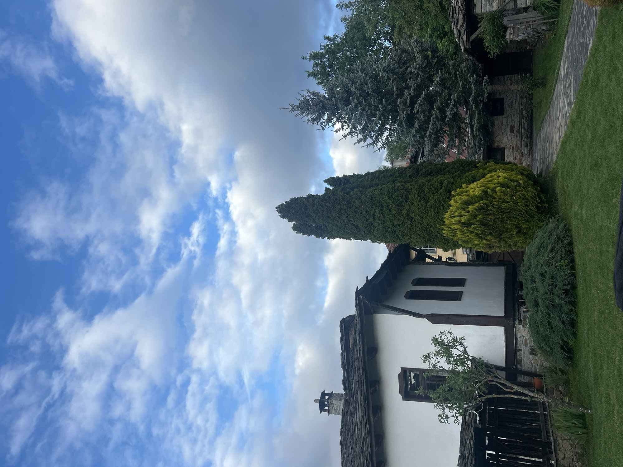 The image depicts a building in the city of Dolen, Bulgaria, under a partly cloudy sky. The building is two stories tall with white walls and dark-colored trim around windows and roof. A stone chimney extends from a dark, tiled roof. Trees and bushes surround the building on the right side of the image. A stone pathway leads away from the building, bordered by grass. The perspective is from below, looking upward toward the building and the sky.