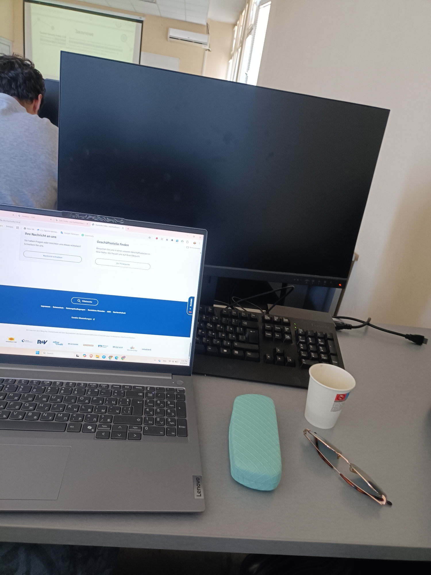 The image shows a workspace with a laptop, external monitor, and keyboard on a grey desk. The laptop screen displays a website in German, possibly related to banking or finance. A white paper cup, a light blue glasses case, and a pair of glasses are also on the desk. The laptop is a Lenovo, with a Cyrillic keyboard. In the background, a person with dark hair is sitting in front of a projector screen. The scene appears to be in an office or classroom setting. An air conditioning unit is visible on the wall.