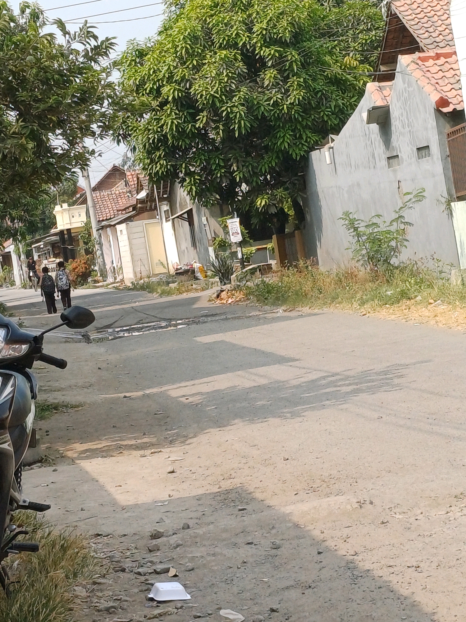 The image captures a residential street in Kersana, Indonesia, bathed in sunlight casting long shadows. In the foreground, a black motorcycle is partially visible on the left side of the frame. The street itself is paved with asphalt and appears somewhat weathered, with patches of discolored material and small puddles. To the right of the road, a gray building with a red-tiled roof is prominent, featuring simple windows and a small protruding awning. Vegetation, including trees with dense green foliage and dry, yellowish grass, lines the sides of the street and the area around the buildings. In the middle ground, two individuals are walking away from the camera down the street, one appearing to wear a backpack. Further down the street, several houses with tiled roofs and visible structures can be seen, interspersed with trees. The sky above is bright and overcast. The overall scene suggests a quiet, everyday moment in a suburban or rural Indonesian setting.
