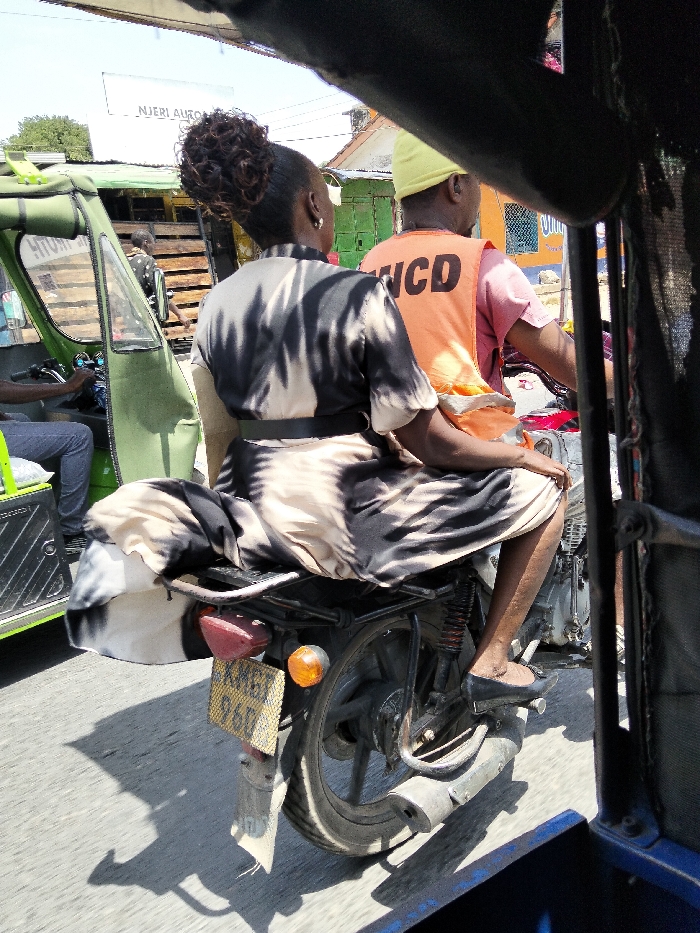 An outdoor street scene in Mkomani ward, Kenya, depicts an adult female passenger riding sidesaddle on the rear of a motorcycle, driven by an adult male. The woman, viewed from behind, has dark hair styled in a bun and wears a black and beige/white patterned short-sleeved dress with a black belt, and black flat shoes. The male driver, also viewed from behind, wears a yellow cap, a pink short-sleeved shirt, and an orange high-visibility vest with "NCD" visible on the back.
The motorcycle is dark-colored, featuring a visible rear wheel, exhaust pipe, suspension, a red taillight, and an amber turn signal. A yellow Kenyan license plate, partially obscured but showing "KMG" and "96D", is attached to the rear. To the left of the motorcycle is a green tuk-tuk (auto-rickshaw), with its driver partially visible inside. The image is framed from within another vehicle, likely a dark-colored tuk-tuk, evident by the dark structure in the foreground.
The environment is an urban setting under bright daylight, indicated by sharp shadows on the paved road. Background elements include various single-story buildings with corrugated metal roofs, some painted in bright colors like green and blue. A white sign with "NJERI AUTO" is visible above the green tuk-tuk. Both the motorcycle and the green tuk-tuk appear to be either in motion or paused in traffic.