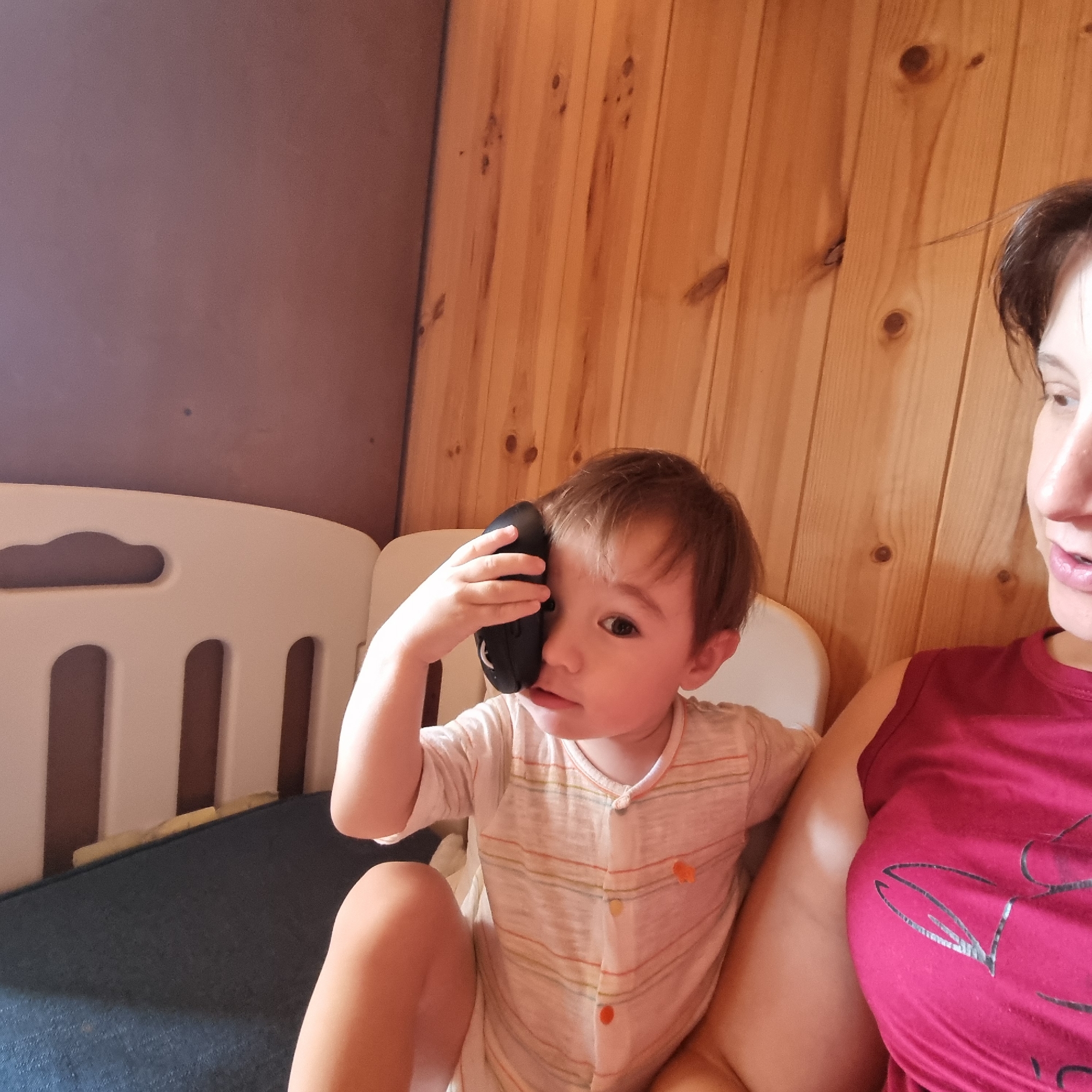 In this indoor, close-up shot, a young child is holding a black computer mouse to the side of their face, covering one eye. The child has light brown hair and is wearing a striped onesie. To the right of the child is an adult woman, partially visible from the chest up, wearing a dark pink t-shirt with a design on it. The background consists of a wooden paneled wall and a white plastic baby gate. The scene appears to be in a domestic setting. The lighting suggests it could be daytime, and there are no strong indicators of weather. The child's expression is one of curiosity or playfulness. There is no visible text in the image.