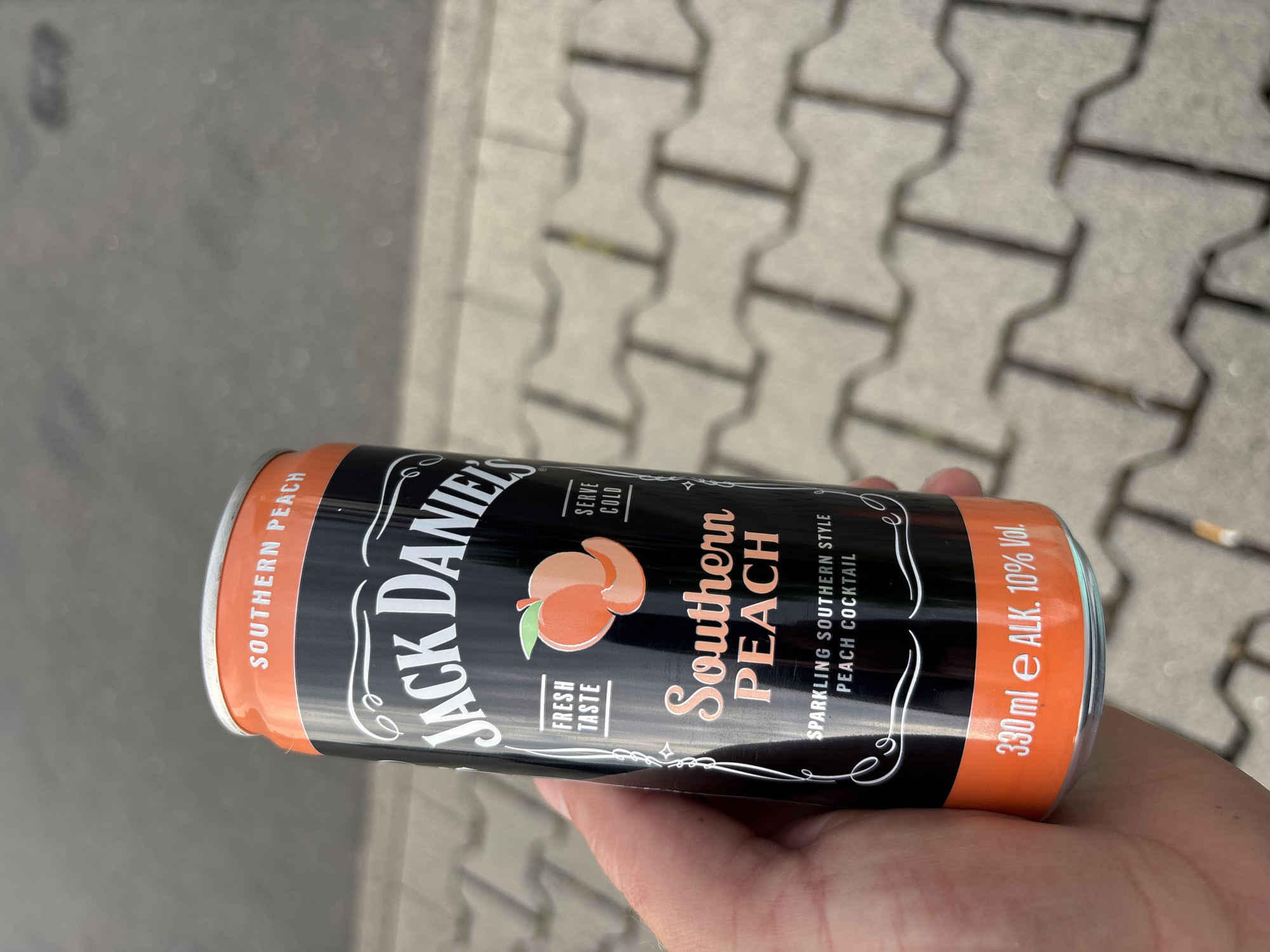 A person holds a black and orange can of Jack Daniel's Southern Peach sparkling cocktail. The can is oriented vertically, with the top facing upwards. The label features the Jack Daniel's logo in white, with "Southern Peach" written in a stylized font below an illustration of two peaches. Text on the can indicates "Sparkling Southern Style Peach Cocktail," "Serve Cold," "Fresh Taste," and "330 ml e ALK. 10% Vol." The background shows a textured grey surface, possibly a paved walkway with interlocking concrete stones. A discarded cigarette butt is visible in the upper right corner of the frame. The lighting suggests it is daytime, outdoors. The image appears to be taken in Elsenfeld, Germany.