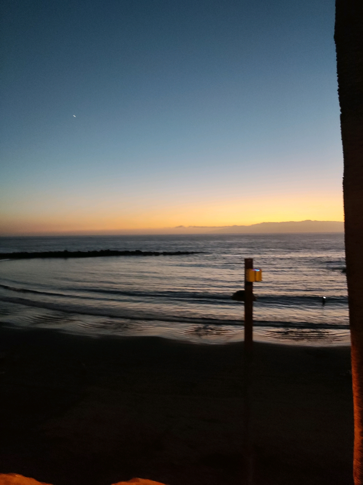 This image depicts a twilight scene on a beach in Costa Adeje, Spain. The sky transitions from a deep blue overhead to a soft orange and yellow hue near the horizon, indicating either sunrise or sunset. The calm ocean water reflects the changing colors of the sky. In the foreground, the dark silhouette of a sandy beach and a palm tree trunk are visible on the right. A small, dark structure with a yellow sign is partially visible in the middle ground, standing in the shallow water or on the wet sand. A faint crescent moon is visible in the upper left portion of the sky. A low, dark breakwater made of rocks extends horizontally across the water in the middle ground. The overall atmosphere is serene and tranquil.