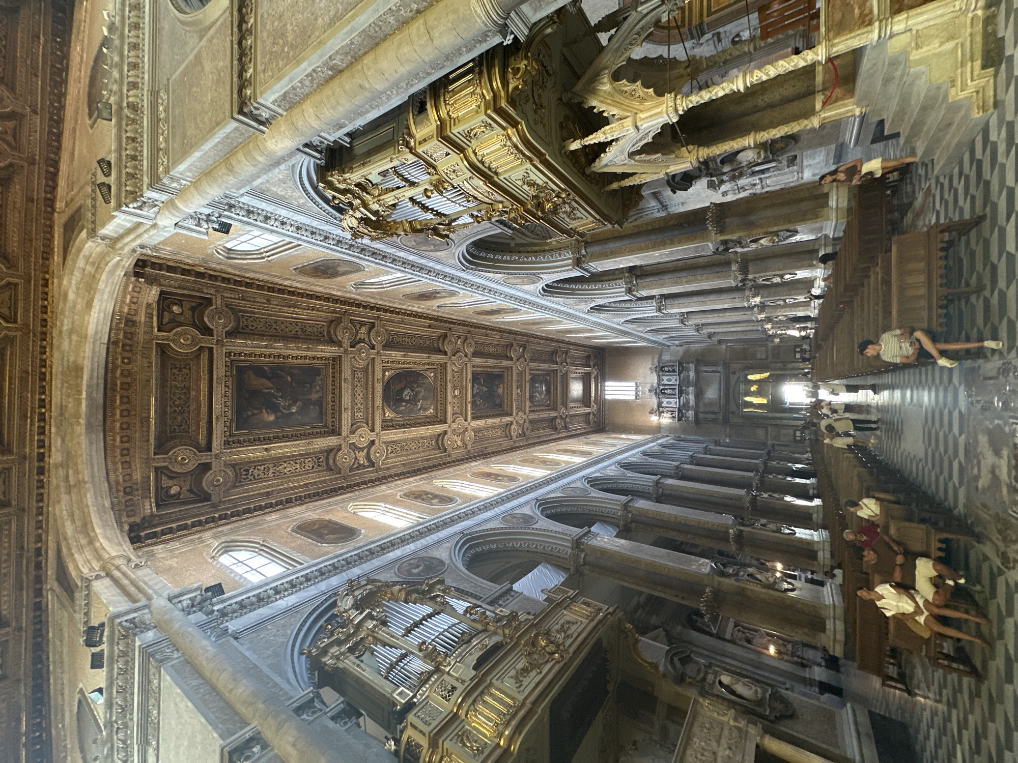 The image depicts the interior of the Naples Cathedral in Naples, Italy. It is a wide-angle shot looking down the nave towards the altar. The cathedral features a high, ornate coffered ceiling with religious paintings. Two large, gilded pipe organs are prominently displayed on opposite sides of the nave, mounted on balconies above arched colonnades. Rows of wooden pews are arranged on either side of the nave, separated by a patterned checkered floor. Several people are visible, some sitting on the pews, while others are standing and walking. The lighting suggests it is daytime, with sunlight streaming through high windows, illuminating the opulent interior. The architecture is predominantly Baroque, characterized by its grandeur and elaborate decoration.