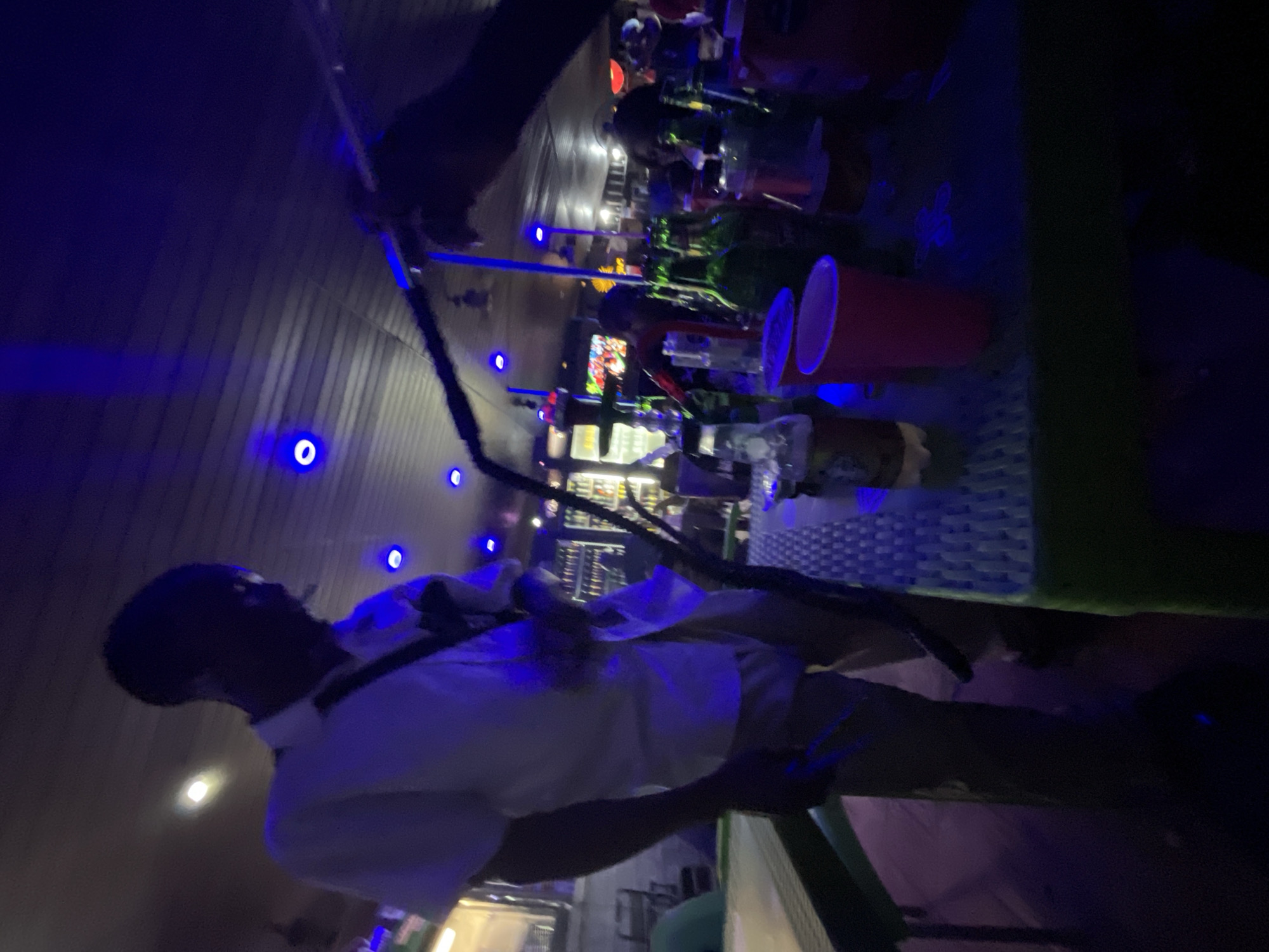 The image depicts a dimly lit indoor setting, likely a bar or club, with a strong blue ambient light casting a glow on the surfaces. A person, silhouetted against the light, is visible from the waist up, wearing a white short-sleeved shirt and a dark strap across their chest. Their face is obscured by shadow. In the foreground, a table is laden with various items, including several glass bottles, some of which appear to be beer bottles, and red disposable cups. A hookah pipe with a black hose is prominently featured, with its stem extending upwards and a hand holding part of the hose. The table surface is textured with a grid pattern. In the background, a bar area is visible with shelves stocked with bottles and illuminated by neon lights. Other people are present in the background, though their features are indistinct due to the low lighting. The overall atmosphere suggests a social gathering. The scene is located in Ado, Nigeria.