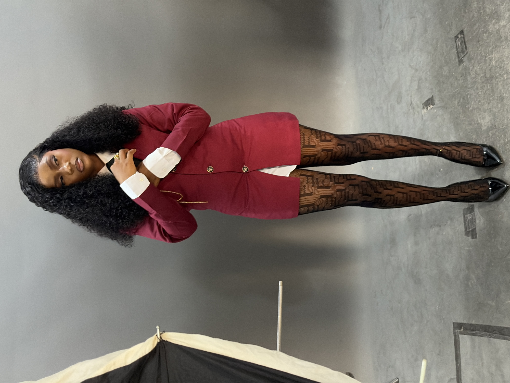 A young woman stands centrally within an indoor studio setting with a plain, medium-grey background and a concrete floor. She has dark, curly hair styled with a part and volume. She is wearing a maroon blazer-style dress with gold buttons, layered over a white collared shirt with prominent white cuffs and a dark tie or cravat. A thin gold chain is visible hanging from the right side of her blazer. On her left hand, she wears a gold ring. Her legs are clad in sheer black tights with a repeating geometric pattern, and she wears black pointed-toe high heels. Her head is turned slightly to her right, and her gaze is directed off-frame to the right. Her hands are positioned in front of her chest, appearing to adjust her right shirt cuff or collar. To the left of the frame, a partially open studio softbox, black and off-white in color, is visible on a stand, with a horizontal metal rod extending from its structure. The concrete floor shows various scuff marks and several dark rectangular outlines.