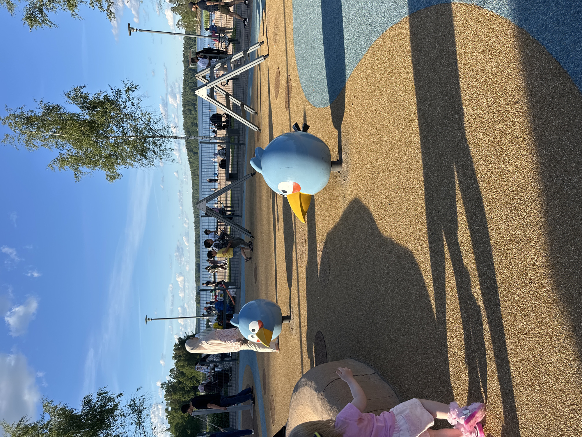 In an outdoor public space, possibly a playground or park area by a body of water, two blue bird-shaped spring riders are prominently featured on a tan and blue rubberized surface. The bird on the left is positioned closer to the foreground and partially obscured by a large, light brown, rock-like structure. The second bird, to the right and slightly further back, is in profile. A young child with blonde hair, wearing a pink shirt and white shorts, stands to the left of the frame, facing away from the viewer and toward the rock. In the background, beyond a metal fence, a body of water stretches towards a tree-lined shore under a bright blue sky with scattered white clouds. Several people are visible in the middle ground, some walking, some sitting, and a few children are engaged in play near playground equipment, which includes two triangular climbing structures. The sun casts long shadows across the playground surface.