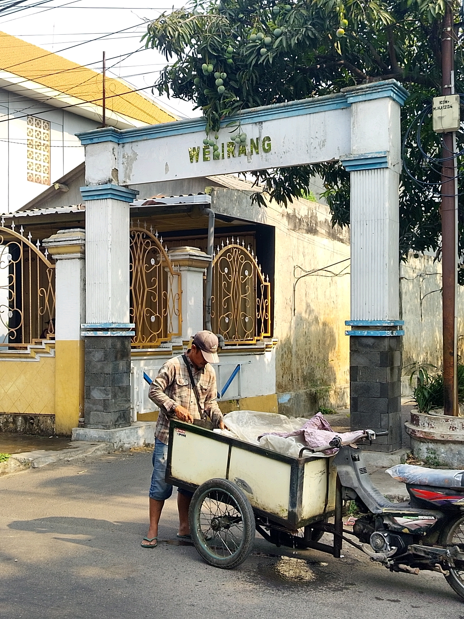 The image shows a man with a tan baseball cap and a plaid shirt standing next to a three-wheeled cart attached to a motorcycle. The cart is partially covered with a white tarp, and there are some fabric items on top of the tarp. The man appears to be attending to the cart. Behind him, there is an archway with the word "WELIRANG" written on it in gold letters. The archway is supported by columns. To the left of the archway is a building with yellow and white walls and decorative metal fencing. To the right of the archway is a utility pole. The scene is outdoors, on a paved street, with trees and vegetation visible. The presence of the "WELIRANG" signage suggests this is likely an entrance or landmark in the city of Mojokerto, Indonesia. The lighting indicates it is daytime.