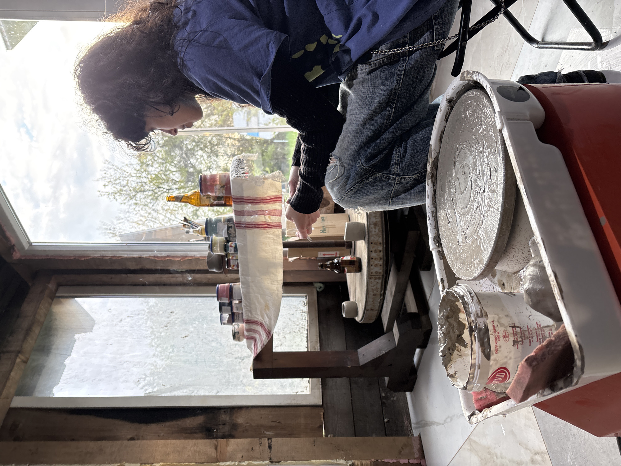 A person with long, dark curly hair, wearing a blue shirt, black long-sleeved shirt, and ripped blue jeans with a chain, is seated at a pottery wheel. They are using their hands to shape a piece of clay on the wheel. A variety of bottles and jars are lined up on a shelf behind them, covered with a white towel with red stripes. There is a broken window to their left, and the room appears to be a workshop or studio. A bucket filled with gray clay sits near the pottery wheel. The location is unknown, but there is no information in the image that says it is in Verinsko, Bulgaria.