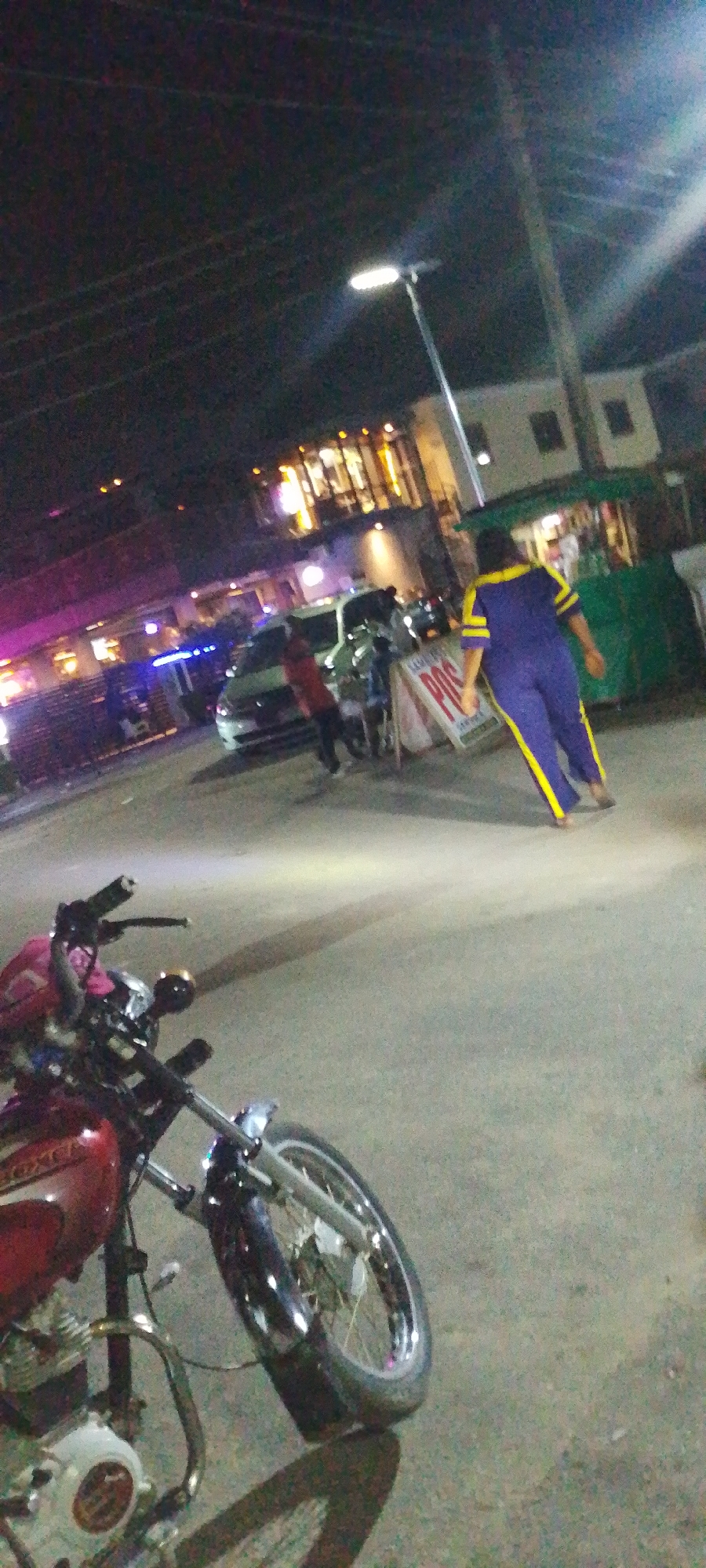 Captured at night in Lokoja, Nigeria, the image presents a bustling street scene. A red motorcycle dominates the foreground, parked on the asphalt. Behind it, a white vehicle is parked in front of a brightly lit building. People are visible in the scene, including one wearing a blue tracksuit with yellow stripes walking toward a green kiosk. The environment includes streetlights, buildings, and a portable sign.
