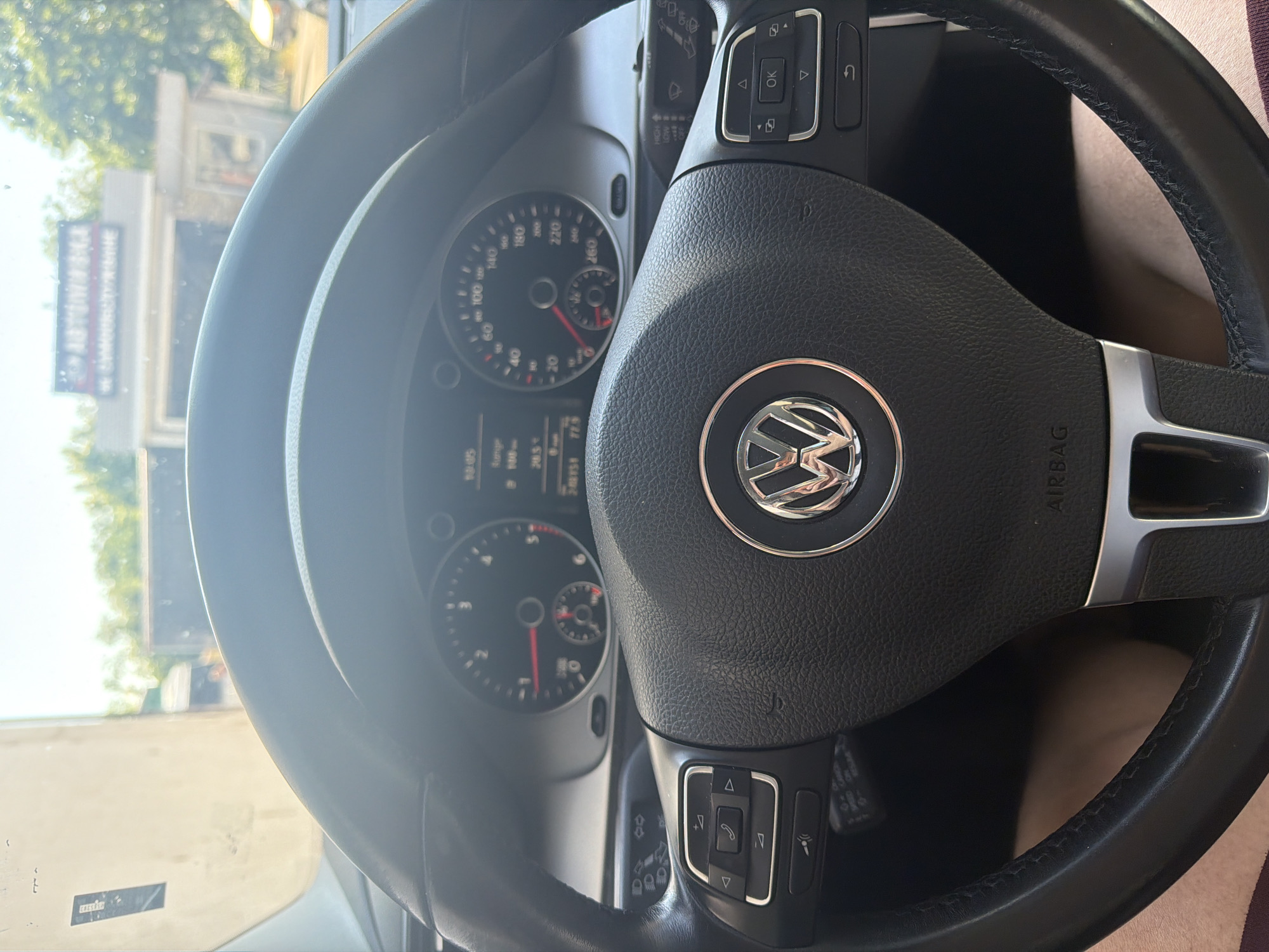 A slightly rotated eye-level shot from inside a car in Shumen, Bulgaria, primarily focuses on the black multi-function steering wheel and a portion of the instrument cluster. The steering wheel prominently features a chrome Volkswagen logo in the center. It has controls on both sides; the right side includes buttons for phone calls and voice commands, while the left side has directional arrows and an "OK" button, likely for navigating the car's display menus. The "AIRBAG" label is visible on the steering wheel's lower right spoke.
Beyond the steering wheel, the instrument cluster is visible, showing a speedometer on the right, marking up to 260 km/h, and a tachometer on the left, up to 7,000 RPM. A digital display between the main gauges shows "19:45" (time), "Runge," "205.1," and "20.5°C," likely indicating the time, trip distance, and outside temperature. Above the steering column, a stalk with wiper controls (HIGH, LOW, OFF) is visible.
Through the windshield, a building with a blue and white sign, partially obscured but appearing to read "AUTOSERVICE" or similar, is visible. Trees are also seen in the background, suggesting an outdoor, urban setting during daylight hours, given the bright illumination. The car appears to be stationary, possibly parked.
