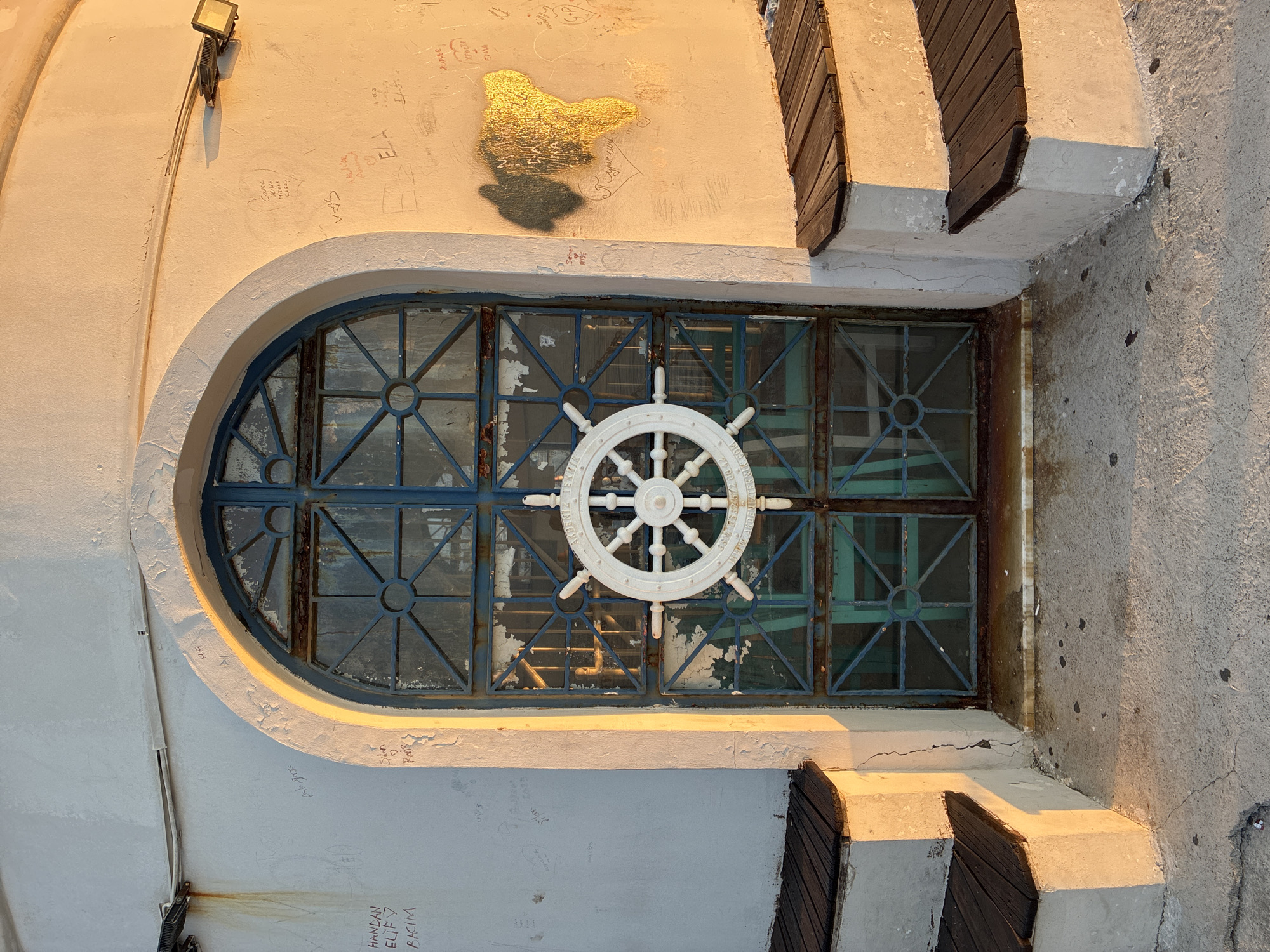 The image depicts the entrance to a structure, possibly a lighthouse or a historical building, in Alanya, Turkey. The entrance is an arched, weathered blue metal door with a white ship's wheel mounted in the center. The door is set within a textured, off-white wall that shows signs of aging and has various graffiti scribbled on it. To the left and right of the entrance are tiered, wooden benches built into concrete steps, suggesting a public area. The scene is illuminated by the warm, golden light of what appears to be sunset, casting long shadows and highlighting the textures of the building and its surroundings. The ground in front of the entrance is paved with concrete, with some cracks and debris visible. A floodlight is mounted on the upper right portion of the wall, aimed downwards.