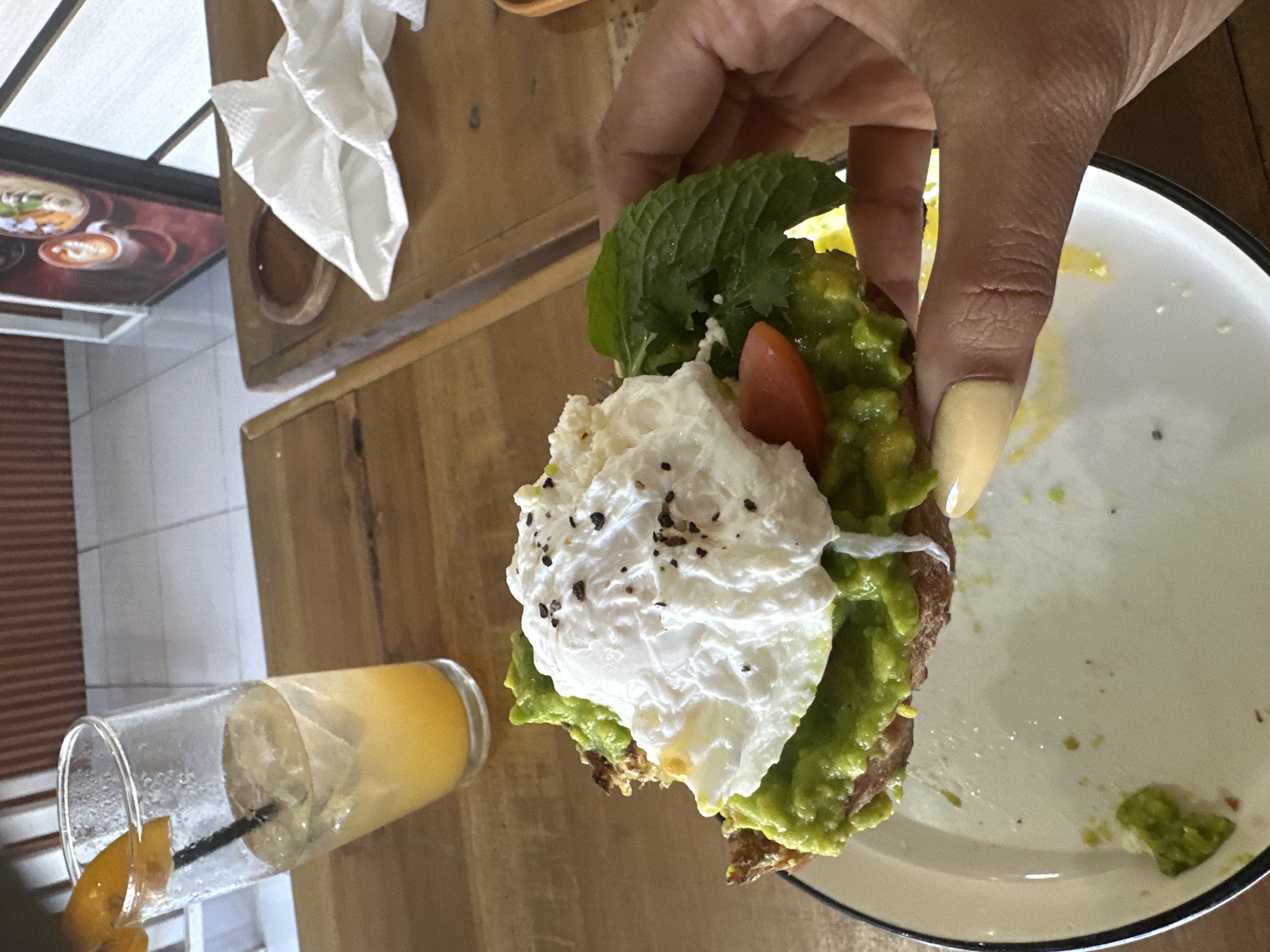 In Kuta Selatan, Indonesia, a close-up shot captures a hand holding a piece of avocado toast topped with a poached egg. The toast is served on a white plate, with avocado spread generously over the toasted bread. A poached egg, perfectly cooked with a runny yolk, sits atop the avocado, sprinkled with black pepper. A slice of tomato and a sprig of mint or cilantro garnish the dish. In the background, a glass of orange juice with ice and a straw is visible, along with a white napkin and a framed picture on a wooden table. The environment appears to be a cafe or restaurant setting.