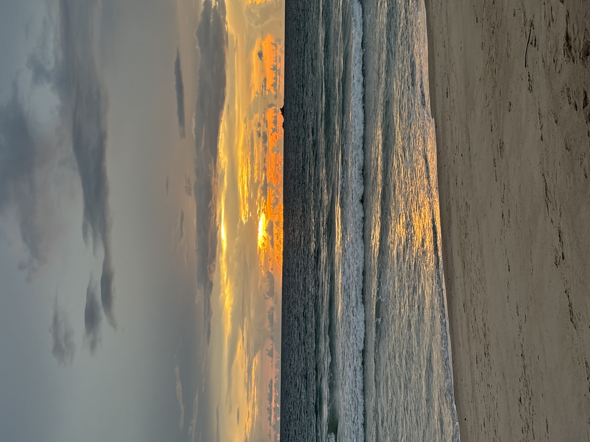 A wide shot captures a beach scene at dusk. The sky is a gradient of grey clouds at the top, transitioning to vibrant orange and yellow hues near the horizon, suggesting a sunset. Darker clouds are present higher up, while lighter, scattered clouds are visible lower in the sky. A thin, dark horizontal line on the horizon appears to be a small island or landmass with some structures. The ocean is a deep blue-green, with moderate waves breaking on the shore, leaving white foam. The wet sand of the beach in the foreground reflects the golden light from the sunset. The beach is mostly empty, with no people visible. The scene is tranquil and atmospheric, typical of a coastal setting in Choeng Thale, Thailand, during sunset.