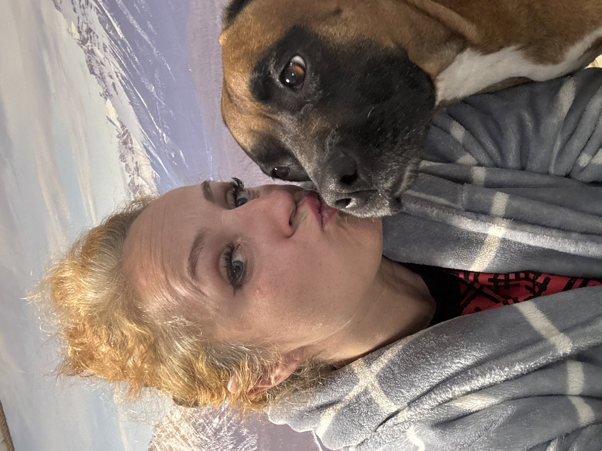 A vertically oriented, close-up portrait captures an affectionate interaction between a woman and a dog. The woman, with fair skin, light blue eyes, and curly blonde hair, occupies the lower left portion of the frame. She looks towards the viewer with pursed lips, forming a "kissy face." She is wrapped in a soft, gray plaid blanket or robe, under which a hint of a red and black patterned garment is visible.
Above her, nestled close, is a medium-sized dog, likely a Boxer or similar breed, with short brown fur and a distinctive black muzzle. The dog's nose is positioned near the woman's lips, and its left eye, with a brown iris, looks directly at the camera. A white patch of fur is visible on the dog's chest.
The background features what appears to be a large wall mural or tapestry depicting a snowy mountain range under a light blue sky, suggesting a scenic and possibly cold environment. The overall emotion conveyed is one of warmth and connection between the woman and her canine companion. No text is visible in the image. The location context provided is Chaikovsky, Russia.