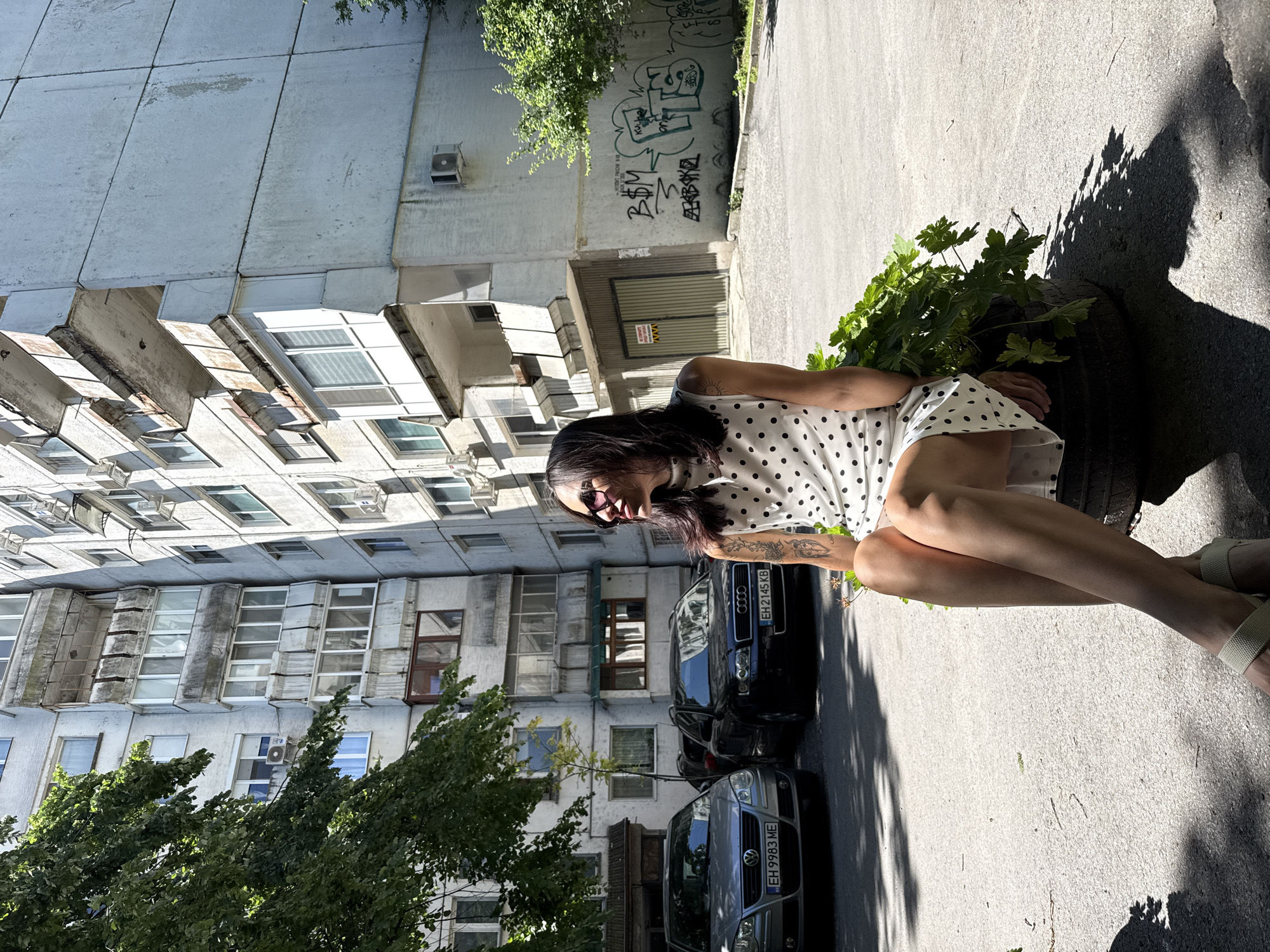 A medium, eye-level, outdoor shot depicts a woman seated in what looks to be a flower planter made from a car tire in Pleven, Bulgaria.
The woman, presumably in her late 20s to early 40s, is seated with her knees to her chest and arms wrapped around her legs. She is wearing a white dress with black polka dots and tan sandals. Her long, dark hair is loose, and she is wearing sunglasses. A tattoo is visible on her left arm.
Behind the woman are two parked cars, one dark and one light gray. The license plates are visible on both cars and read "EH 2145 KB" and "EH 9983 ME". Behind the cars is a large, concrete building with many windows. Graffiti can be seen on a part of the building to the right of the woman. The building appears to be a multi-story apartment complex, indicative of urban residential architecture. Trees can be seen to the left of the building, casting a shadow on the ground. The ground is paved and appears to be part of a street or parking area.
