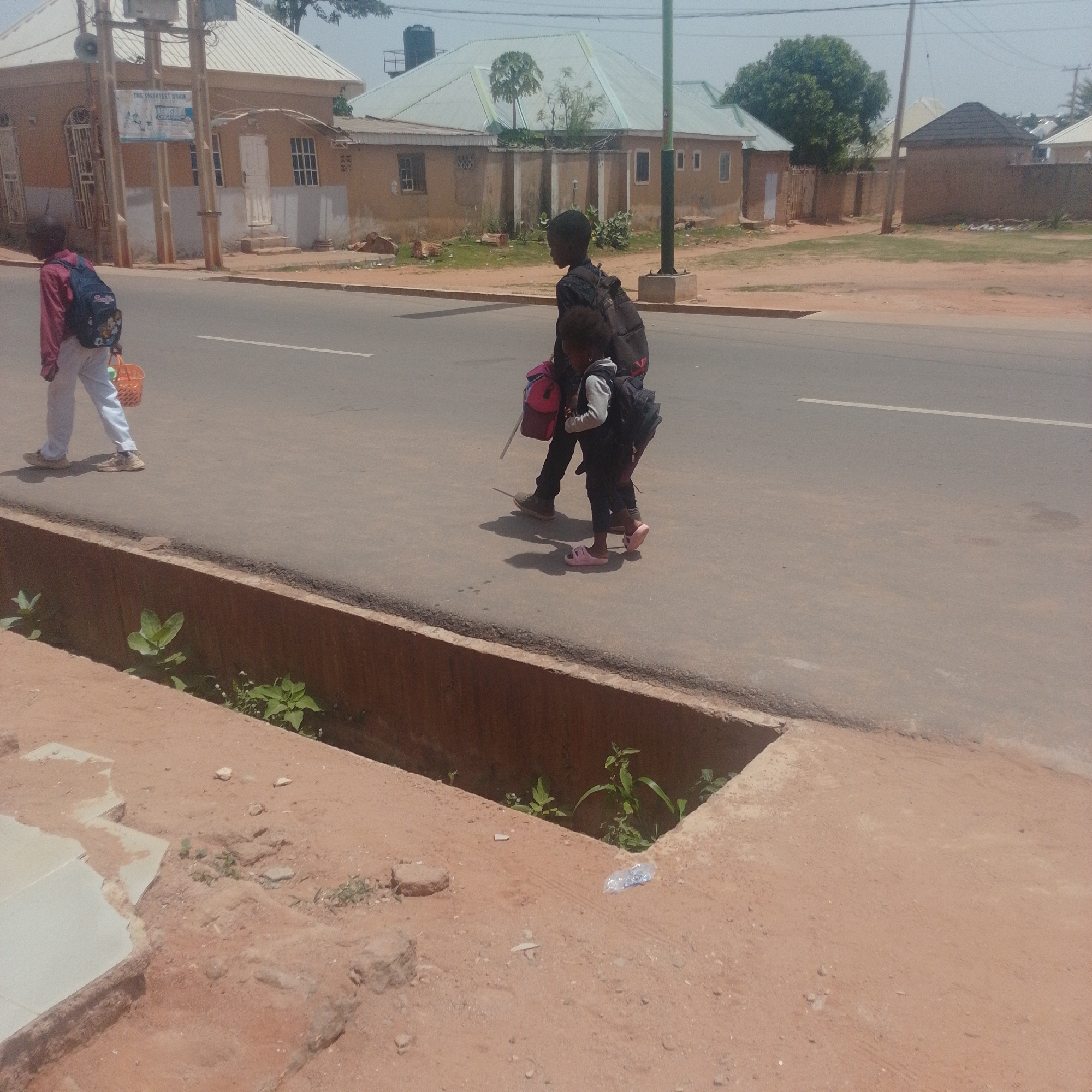 In Tempure, Nigeria, two children walk along a paved road under a clear sky. The older child, on the left, wears a reddish-pink long-sleeved shirt, white pants, and sneakers, carrying a blue backpack and a small orange basket in their left hand. The younger child, on the right, walks behind the older child, wearing dark pants, a gray sweater, and pink sandals, carrying a black backpack. They are walking on the right side of the road, with a concrete drainage ditch alongside the road. Buildings with tan walls and light-colored roofs are visible in the background. The environment appears dry, with patches of vegetation growing in the drainage ditch and sparse grass on the side of the road.