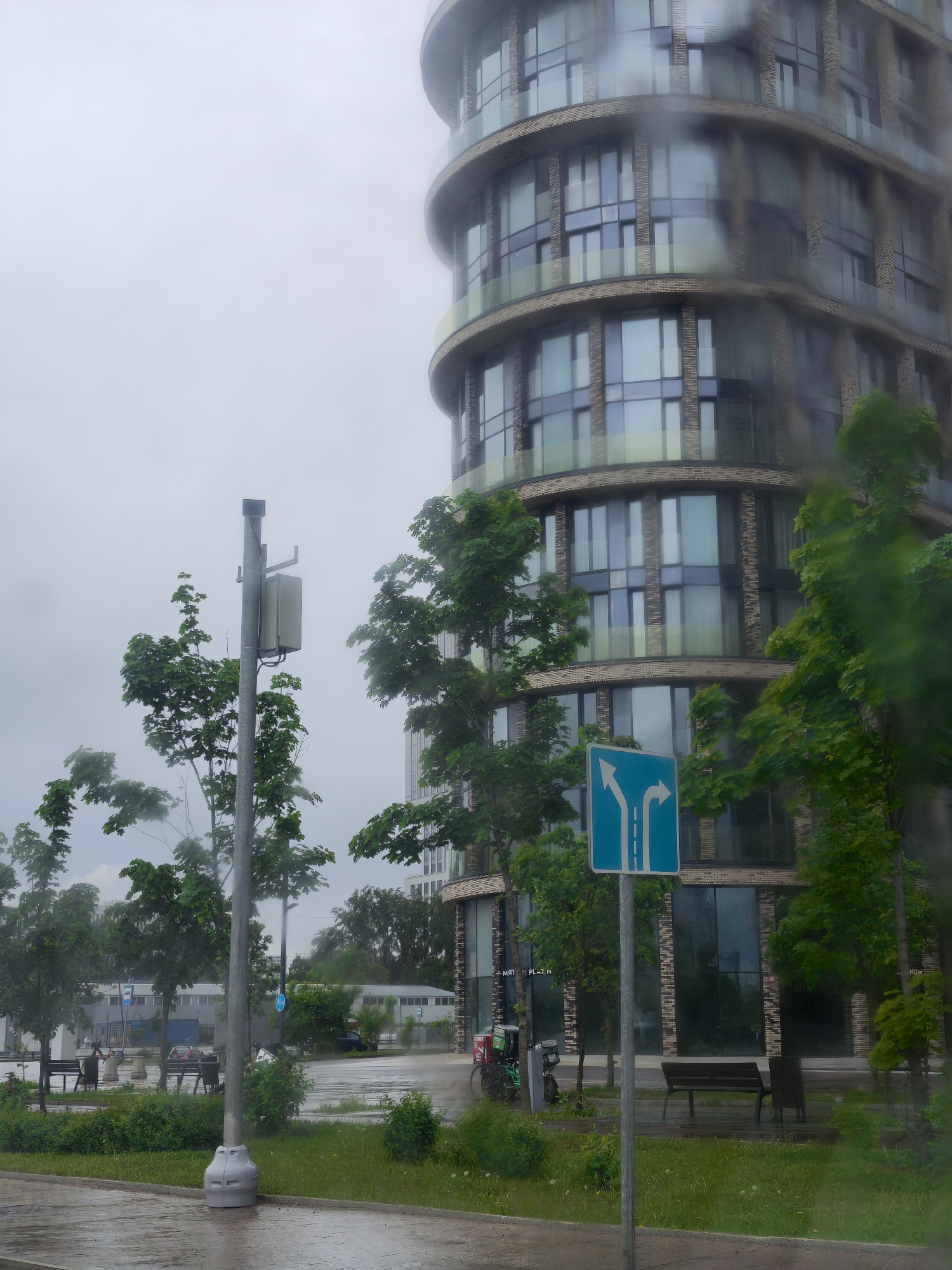 The image depicts a street scene on a rainy day in Moscow, Russia. The foreground shows a wet pavement with puddles, indicating recent rainfall. A traffic sign with blue and white arrows pointing left and right is prominently placed on the right side of the frame. The sign is surrounded by green trees and foliage. A modern, cylindrical building with rounded balconies and reflective windows rises in the background. The building has a facade composed of brick and glass. Trees are sparsely planted along the street. The street is wet from the rain. Benches are visible, although no people are immediately apparent in the immediate foreground. The atmosphere is overcast with a grey sky. There are parked scooters visible next to the benches.