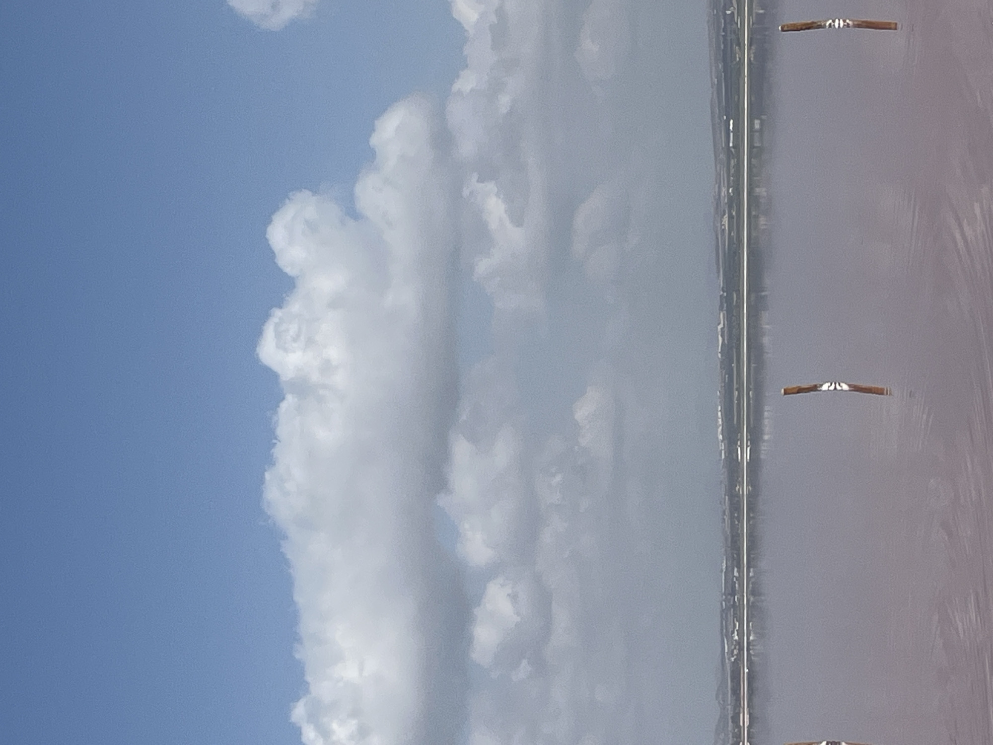 This is a full-frame, eye-level shot of a pink salt lake with a distant shoreline under a cloudy blue sky. Several wooden posts are visible sticking out of the shallow, pink water, suggesting it is part of the salt extraction process at the Salinas y Arenales de San Pedro del Pinatar Natural Park or the nearby Laguna Rosa de Torrevieja, both in the Torrevieja area of Spain. The distant shoreline is low-lying and appears to be lined with buildings and vegetation. The sky is partly cloudy with large, fluffy white clouds dominating the upper portion of the frame. The water's surface is calm with subtle ripples, reflecting the light and the distant landscape. There are no people or animals visible in the image.