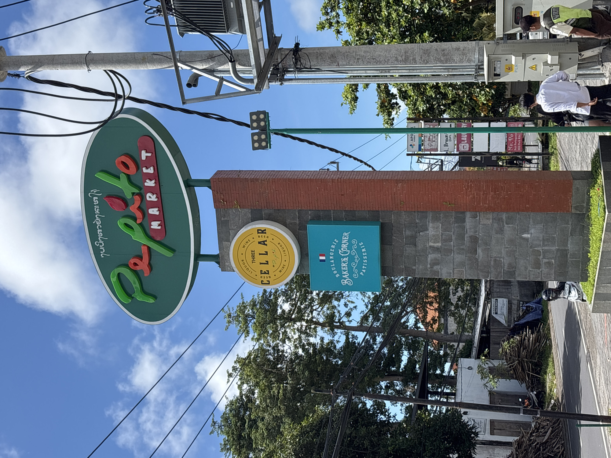 The image shows a street in Ungasan, Indonesia, with a sign for Pepito Market. Adjacent to the Pepito Market sign are two smaller signs advertising "Three Cellar" (wine, beer and liquor) and "Baker's Corner" (boulangerie & patisserie). The signs are mounted on a brick and stone pillar. Power lines are visible in the foreground and a pole is on the left. Further down the street, other business signs are visible including APO Mania, L Brown Store, LAVI, and MP. Po (Best Portuguese BBQ Pork Ribs). Two people can be seen by what seems to be an electrical unit.