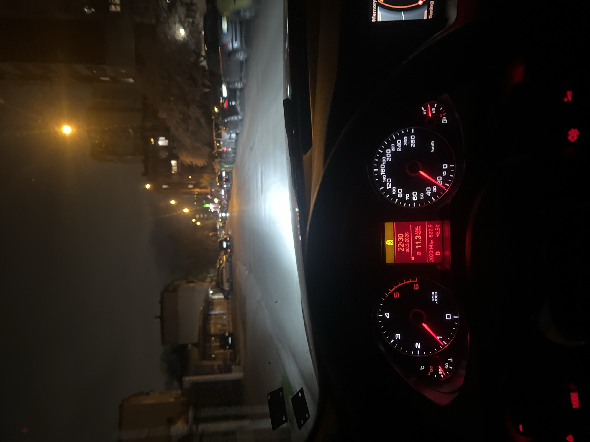 The image is taken from inside a car, likely the driver's seat. The car is moving down a street in what appears to be an urban environment at night. Streetlights illuminate the road and buildings are visible on either side. Other vehicles are also present on the road. The dashboard of the car is visible, displaying information such as the time (22:30 on March 30, 2026), the outside temperature (+6.5 degrees Celsius), the car's speed (approximately 25 km/h), and the mileage (202314 km). Based on the Cyrillic script on the speedometer and the road markings, the location is likely Sofia, Bulgaria.