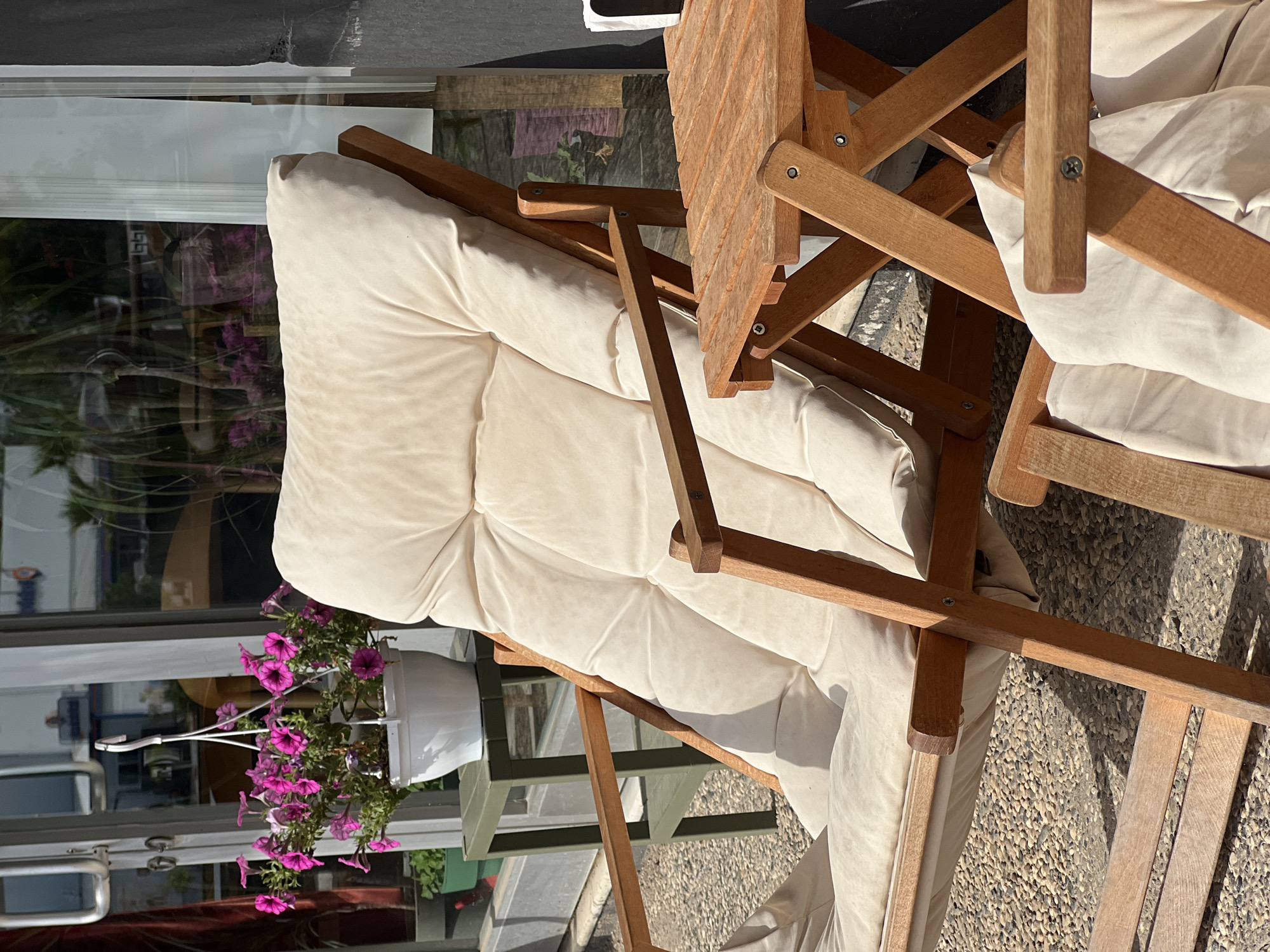 This image depicts an outdoor seating area, likely a cafe or restaurant patio, in Antalya, Turkey. In the foreground, a beige-cushioned wooden lounge chair is prominently featured. To its right, a small wooden folding table is visible. The ground is covered in small, light-colored pebbles. In the background, a glass-fronted building is reflected, showing some greenery and a white hanging pot with pink petunias. The overall impression is one of a relaxed outdoor space bathed in sunlight.