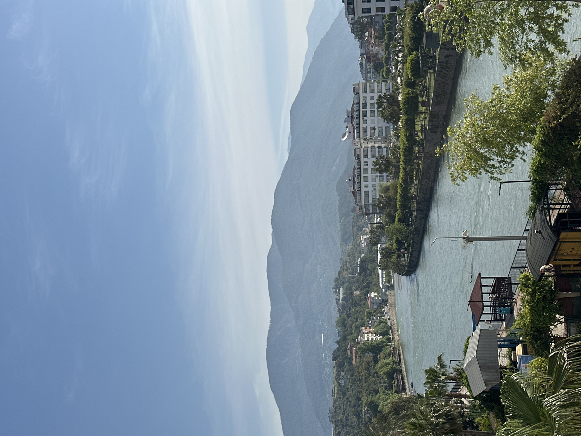 The image presents a scenic landscape photograph, captured in portrait orientation, depicting a town situated between a body of water and large mountains under a clear sky.
Mentally rotating the image 90 degrees counter-clockwise for natural viewing, the scene features a wide body of water, likely a river or canal, flowing from the background towards the foreground on the right. The water's surface is gently rippled, reflecting bright daylight. Along the far bank, a promenade or pathway is visible, lined with trees and railings. On the near bank, a lamppost, a covered seating area with a reddish roof, and a small building are discernible, surrounded by rich green foliage and palm trees.
To the left of the waterway, extending up the slopes and into the mid-ground, are numerous multi-story residential buildings. These light-colored, modern structures are nestled among dense green trees and vegetation. A prominent white building with balconies and a satellite dish is clearly visible among them.
Dominating the background and spanning the left side of the frame are expansive, undulating mountains. Their peaks appear somewhat hazy, indicating atmospheric distance. Above the mountains and across the upper left of the image, the sky is a bright, clear blue with soft, wispy clouds.
The scene depicts a sunny day, likely mid-morning to late afternoon, characterized by strong, natural lighting. No people are visible, and no specific activity is taking place, presenting a peaceful, static landscape. The combination of mountains, water, and developed areas is consistent with the provided location context of Alanya, Turkey. There is no visible text in the image.