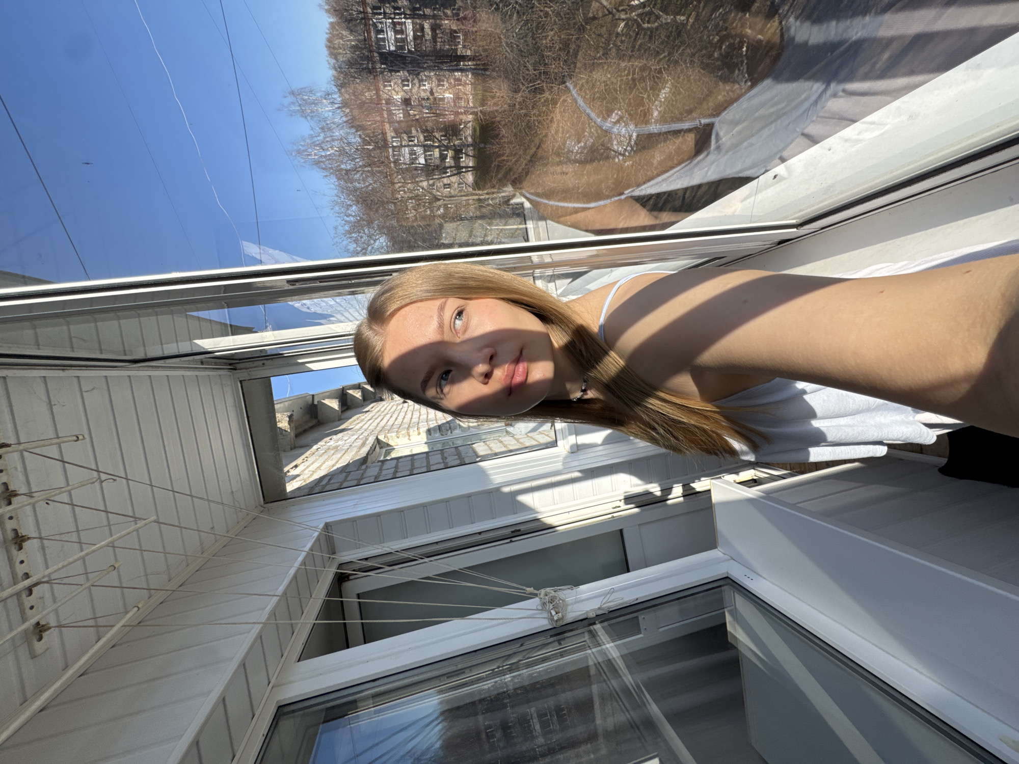 The image captures a brightly lit, top-down selfie of a young woman within an enclosed balcony or sunroom. The woman, with long, straight blonde hair and a fair complexion, looks calmly towards the camera. Her face is bisected by a strong shadow from the bright overhead sun, highlighting one side while leaving the other in partial shade, creating a dramatic contrast. She wears a light-colored tank top, with additional shadows cast on her arm and clothing. A thin, delicate necklace is visible around her neck.
The surrounding environment of the balcony consists predominantly of white, modern window frames and large glass panels. These panels reflect a clear, bright blue sky and the distant outdoor scenery, which includes bare-branched trees, indicative of late winter or early spring, and multi-story residential buildings. These reflections provide a glimpse into the urban setting of Chaikovsky, Russia. On the left side of the balcony, a practical clothesline system is mounted on a white paneled wall, featuring several white bars with multiple strings crisscrossing, some of which appear tangled. The walls and possibly the ceiling of the enclosure are made of white, grooved paneling. The strong sunlight bathes the scene, creating sharp lines and bright areas, consistent with a clear daytime. There is no visible text in the image.
