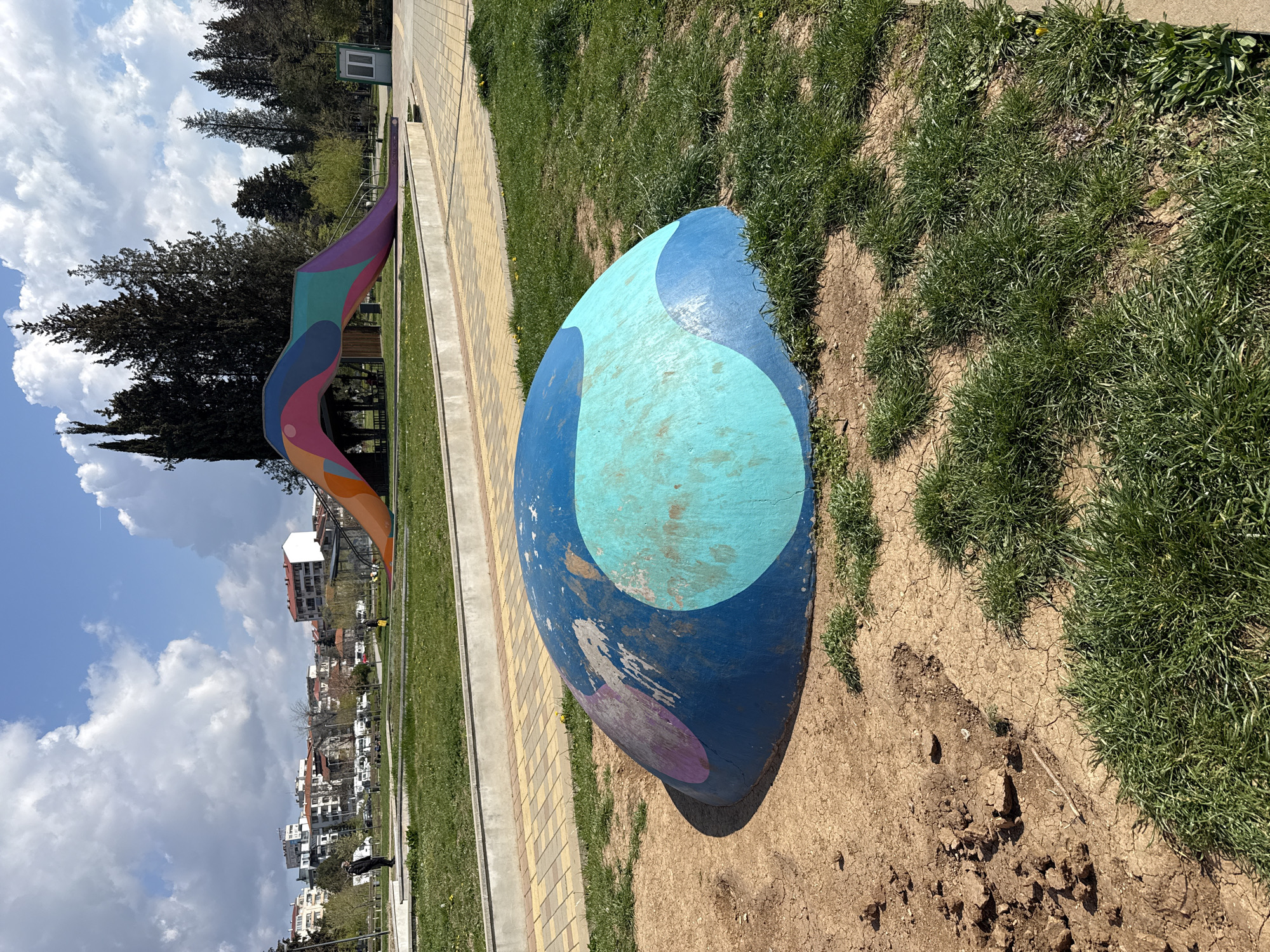 The photograph depicts a public park in Stara Zagora, Bulgaria, showcasing a large, painted rock near a pedestrian walkway. The rock is painted with shades of blue and purple in an abstract design, partially surrounded by grass and bare soil. In the background, there's a colorful, ribbon-like sculpture and residential buildings under a partly cloudy sky. The overall scene captures a sunny day in an urban green space.