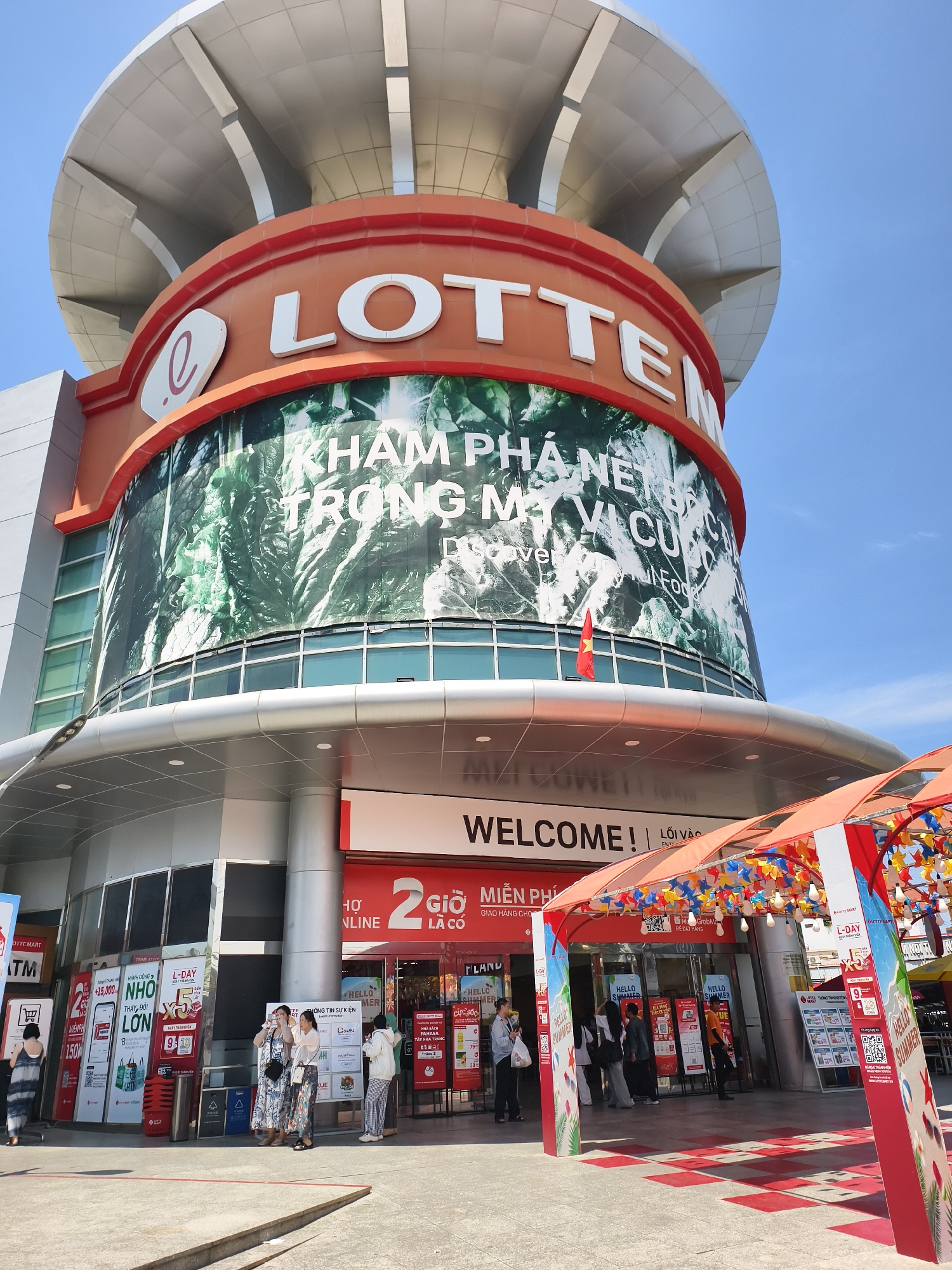 The image displays the exterior of a multi-story commercial building identified as "LOTTE" in Tay Nha Trang Ward, Vietnam, under a clear blue sky. The upper section of the building features a wide, circular, light-colored roof structure composed of overlapping panels. Below this, a large cylindrical facade is painted orange with prominent white block letters spelling "LOTTE" and an associated 'e' logo. Directly beneath the "LOTTE" signage, a large banner wraps around the building, featuring a green, leafy pattern and white Vietnamese text reading "KHÁM PHÁ NỀN ẨM THỰC TRONG MỘT VỊ CỦA," with smaller English text stating "Discover the World of Food." A small red Vietnamese flag with a yellow star is affixed to the building's facade below this banner.
The ground level features a broad entrance area with glass windows and structural columns. Above the main entrance, a white sign reads "WELCOME!" to the left, and Vietnamese text "LỐI VÀO" (Entrance) to the right. Numerous promotional posters and freestanding banners, predominantly in Vietnamese and some displaying "L-DAY," "MIỄN PHÍ" (Free), and "HELLO SUMMER," are positioned near the entry points. An "ATM" sign is visible on the far left. A decorative red and white archway, adorned with colorful pinwheels and string lights, extends from the right side of the entrance area, partially covering the pedestrian path.
Several individuals are present at the entrance. On the left, one person in a light-colored, patterned dress walks away from the building. In the center, a group of four women are standing near the glass entrance doors, some appearing to interact or look at their phones. Other individuals are partially visible further inside the entrance or near the decorative archway. The ground in the foreground is paved, transitioning to a red and white checkered pattern beneath the archway. The scene is illuminated by bright natural sunlight, indicating clear weather.