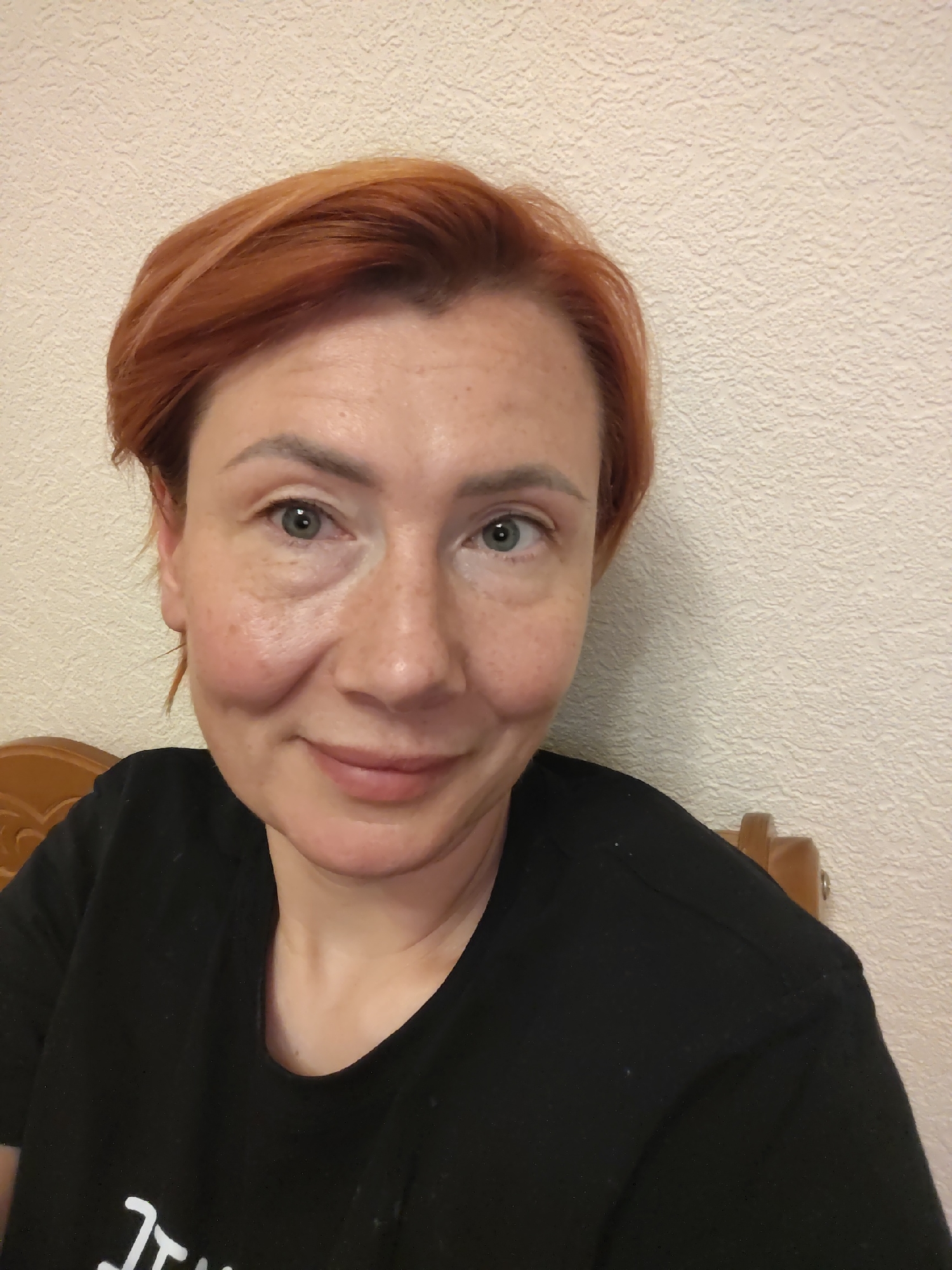 A woman with short, reddish-brown hair and green eyes is looking at the camera. She is wearing a black t-shirt. Her face is slightly angled to the left, and she has a faint smile. The background is a textured, light-colored wall, and a portion of a wooden chair is visible to the left and right of the woman. The lighting is even, with no harsh shadows. There are no discernible elements to indicate the specific location within the city of Konakovo, Russia.