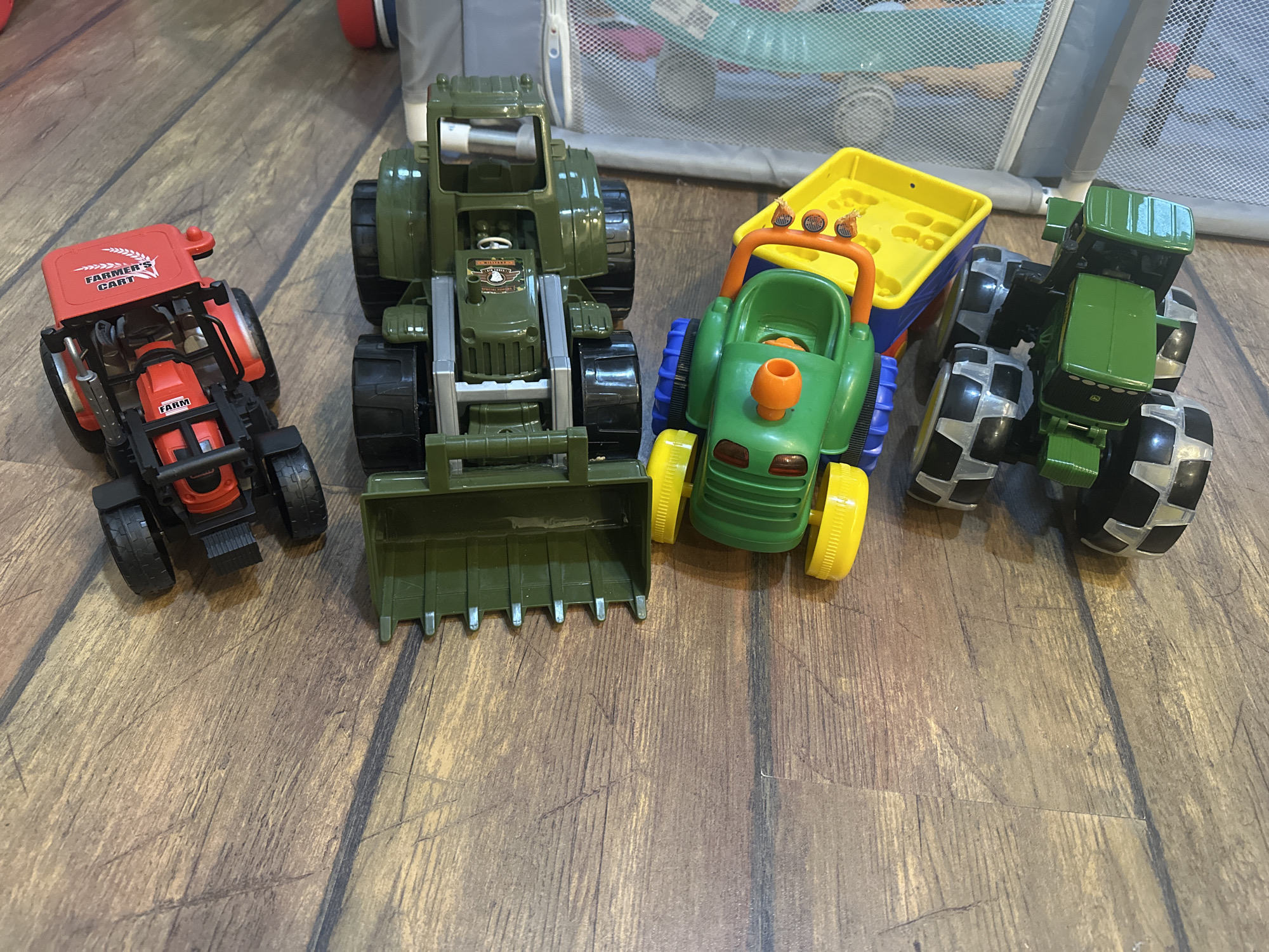 The image depicts a collection of toy farm vehicles on a wooden floor. From left to right, there is a red tractor with the words "FARMER'S CART" printed on its hood, a large green tractor with a front loader, a green and orange tractor with a yellow trailer, and another green tractor. The background shows a portion of a gray, mesh-like structure, possibly a playpen or a storage bin. The lighting suggests an indoor environment, likely a home. There are no people visible in the image. The scene is static, with the toys arranged in a still life composition. The image does not provide any information about the location in the city of Kyiv, Ukraine.