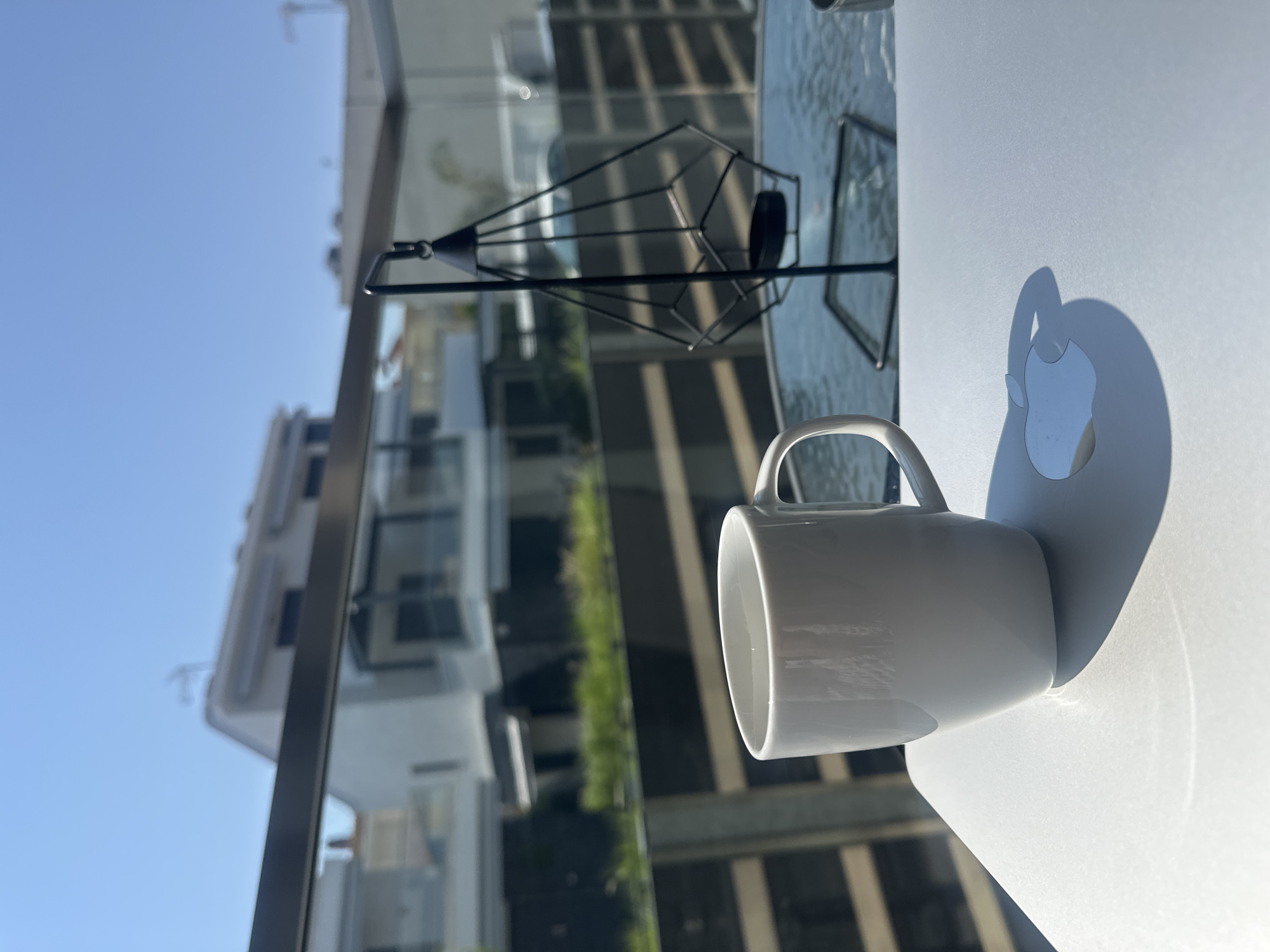 A white coffee mug sits on a silver laptop with an Apple logo. In the background, a modern house with balconies and green foliage is visible under a clear blue sky. To the right of the mug, a black geometric lantern hangs from a stand. The scene is outdoors, likely on a balcony, with bright sunlight casting shadows.