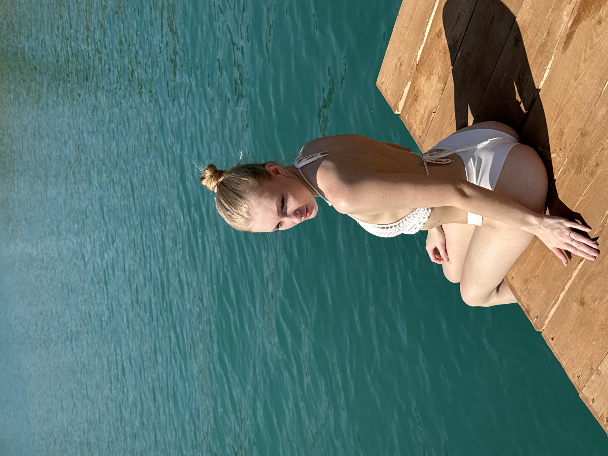 A young woman with blonde hair pulled into a bun sits on a wooden pier overlooking a body of turquoise water. She is wearing a white crochet bikini and looking over her left shoulder. Her hands are resting on her legs. The pier is made of weathered wooden planks and is casting a shadow on the right side of the image. The water appears calm with small ripples. The background is out of focus, suggesting a natural environment.