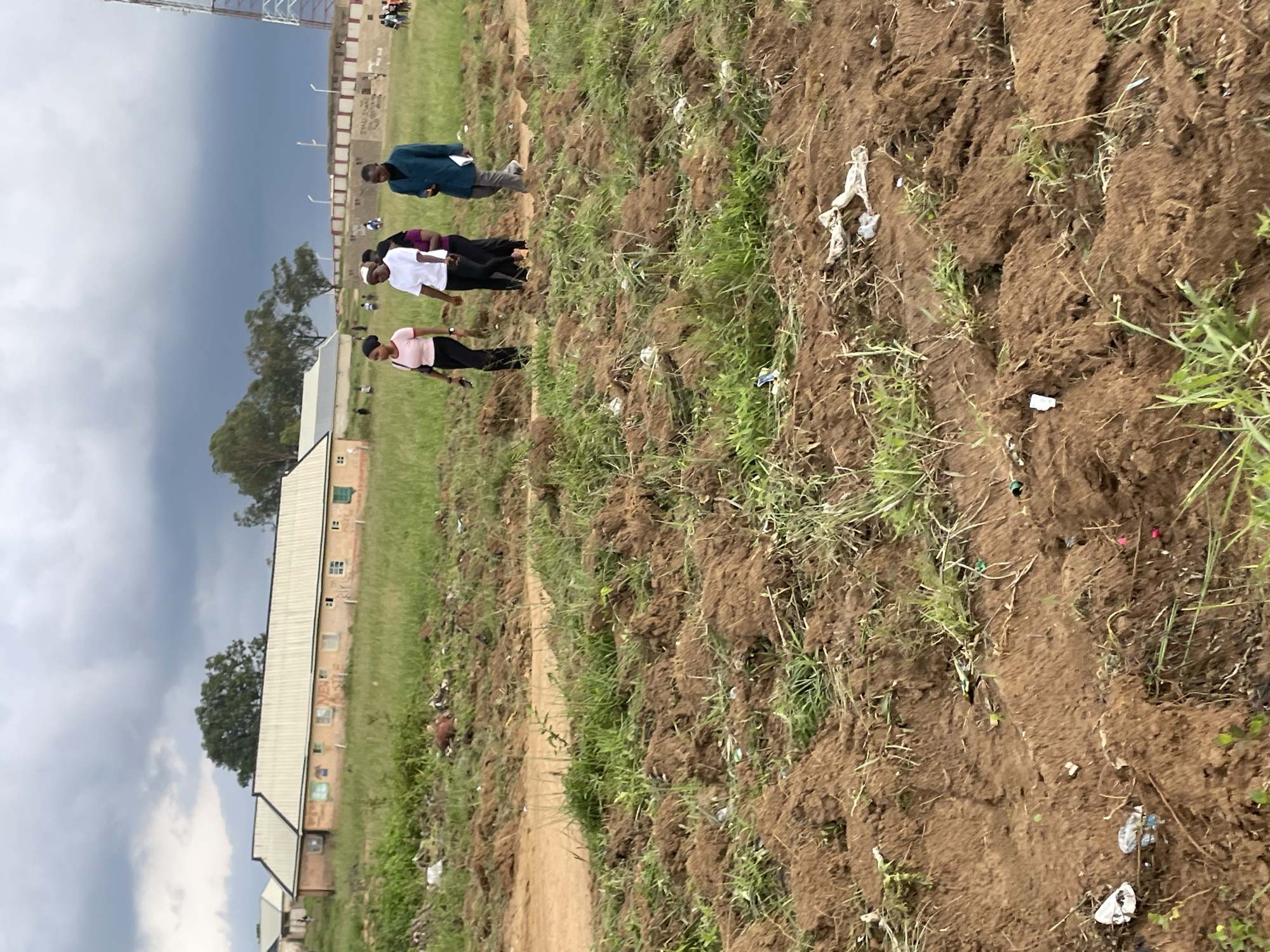 The image shows an overcast sky above a rural or semi-rural environment in Durumi, Nigeria. In the background, there is a low-rise building with a light-colored roof and walls painted a pale orange. The building has multiple windows. Further in the background, a tall telecommunications tower is visible. In the middle ground, a group of four individuals are walking across a grassy area. The person in the foreground on the left is a woman wearing a pink top and black pants, holding a phone. Next to her is another woman in a white top and black pants. Behind them is a third woman in a purple top, and to her right, a man in a teal jacket and gray pants. The foreground is dominated by a patch of tilled, brown earth with sparse green grass growing through it, interspersed with litter, including white plastic bags and other debris. The overall scene suggests an outdoor area, possibly undergoing construction or land preparation, with evidence of human activity and environmental neglect.