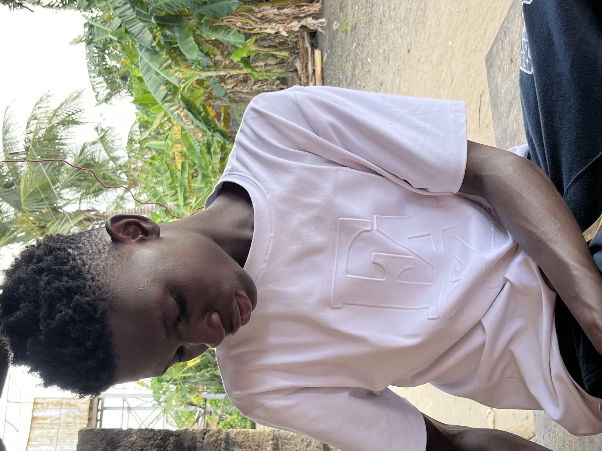 A young black man in a white t-shirt is positioned in the center of the frame, looking down with closed eyes and a relaxed expression. The location is likely in Badagry, Nigeria, indicated by the presence of lush tropical vegetation including banana trees in the background. The man is outdoors, the setting suggests a residential or semi-rural area. He is seated next to someone wearing dark blue clothing, only partially visible on the right. The scene is well-lit, suggesting it is daytime.