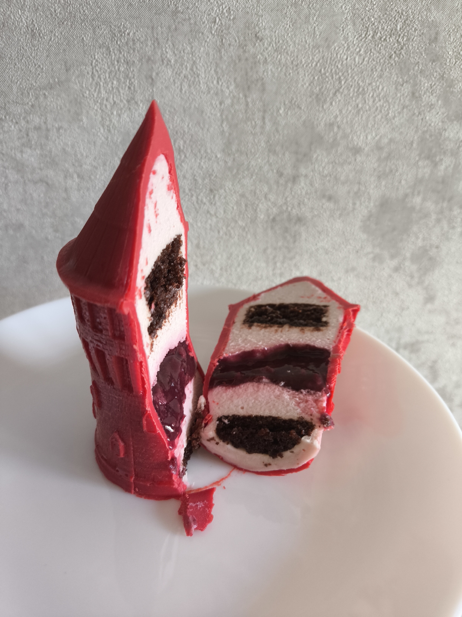 An elaborately crafted dessert, shaped like a vibrant red tower or castle turret, is presented on a clean white plate against a light, textured background. The dessert has been cut longitudinally, revealing its intricate internal layers.
The exterior is a glossy, bright red, possibly a chocolate shell or glaze, molded with details that suggest architectural features like windows or crenellations on its cylindrical base and a pointed roof. One half of the dessert stands upright, showcasing its full design, while the other half lies on the plate, displaying its cross-section.
The interior consists of distinct layers: light pinkish-white cream or mousse, alternating with dark brown cake or brownie sponge. A prominent, thick layer of translucent, dark red or purple fruit jelly, likely cherry or berry, is visible in the middle. The varying textures, from the smooth outer shell to the soft cream, dense cake, and gelatinous fruit filling, are clearly visible.
The image captures the dessert as if it has just been served and cut, ready for consumption. The background is a plain, light grey or off-white wall with a subtle, rough texture and a diagonal woven pattern. No people or visible text are present. The scene is located in Moscow, Russia.