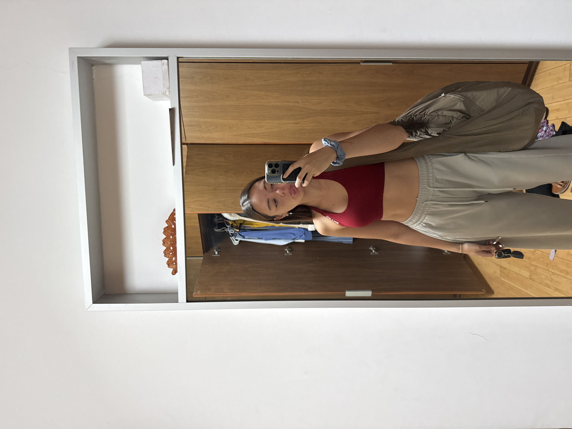 A young woman is taking a selfie in a mirror. She is wearing a red sports bra and light gray sweatpants. She has a blue scrunchie in her hair. The mirror is part of a wardrobe with wooden doors. Open shelves above the wardrobe contain clothing and decorative items. The setting appears to be inside a room with white walls. There are no clues indicating this is San Bernardino Tlaxcalancingo, Mexico.