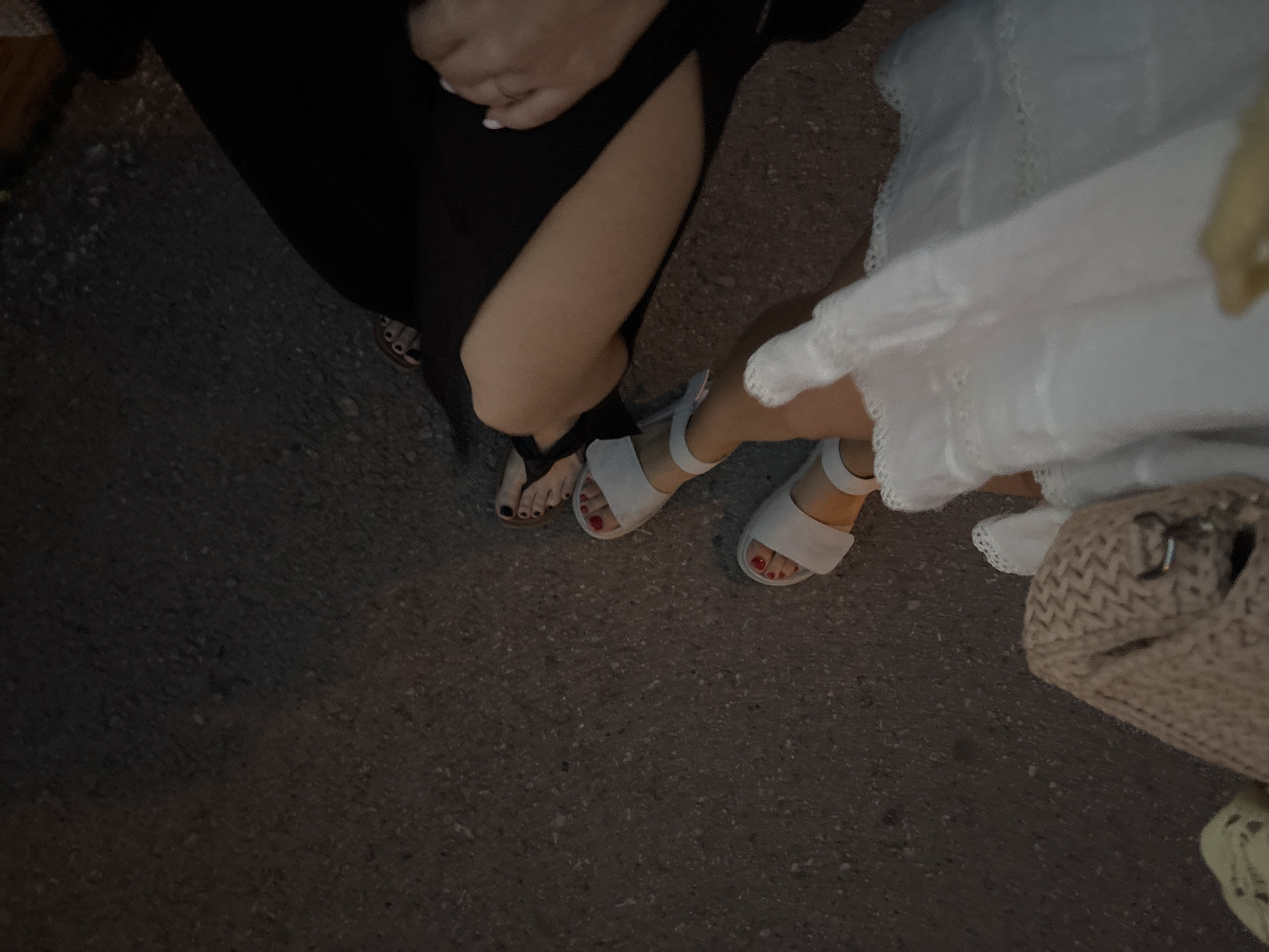 The image shows two individuals standing on a paved surface, presumably a street. The individual on the left is wearing a black dress with a slit, exposing a portion of their leg and foot, which is adorned with a black pedicure. They are wearing sandals. The individual on the right is wearing a white dress and white sandals, with red nail polish on their toes. A beige woven bag is visible near the bottom right corner of the image. The location cannot be determined from the image.