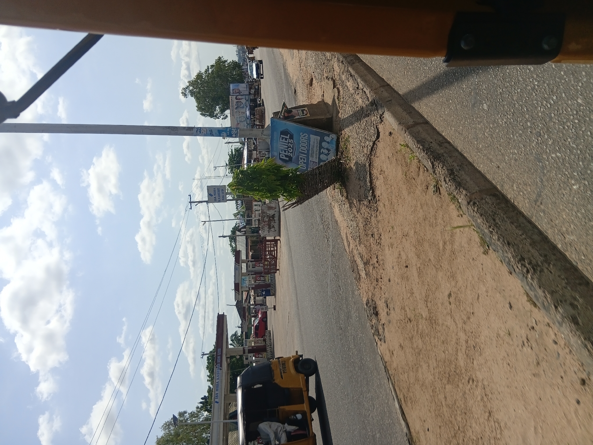 The image depicts a street scene in Jalingo, Nigeria, on a partly cloudy day. A yellow motorized tricycle, known locally as an "achaba," is moving from left to right in the foreground, with a passenger visible in the back. The street is paved, with a sandy shoulder to the right. In the background, a gas station with multiple pumps is visible, identified by signage as "ISA MUSA KASHIME NIG LTD." Further down the road, a white car is driving away from the camera. A large billboard with images of people is also present. A triangular blue and white sign with the text "PENFIEL 2025 OPEN DOORS" and images of faces is propped up on the side of the road, next to a small, decorative tree. Electrical wires are strung overhead, supported by poles. The overall scene suggests a busy urban environment.