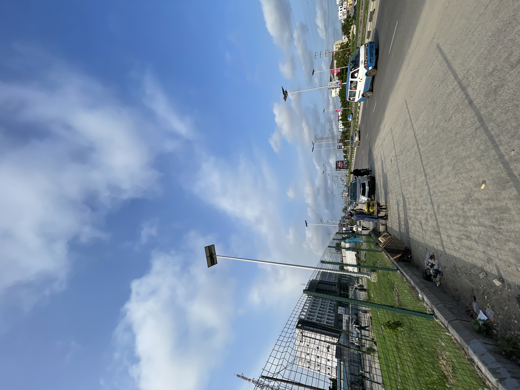 The image depicts a street scene in Lekki, Nigeria, under a partly cloudy blue sky. A multi-lane road occupies the right side of the frame, with several vehicles including a white and blue van, cars, and a motorcycle. On the left, a fenced-off area with a grassy lawn is visible, bordered by a sidewalk. The sidewalk is strewn with litter, including plastic bags and debris. Several solar-powered streetlights line the road. In the background, a large, modern building with numerous windows stands behind the fence. People are seen walking on the sidewalk and near the road, some appearing to be vendors. The overall impression is of an active urban environment.